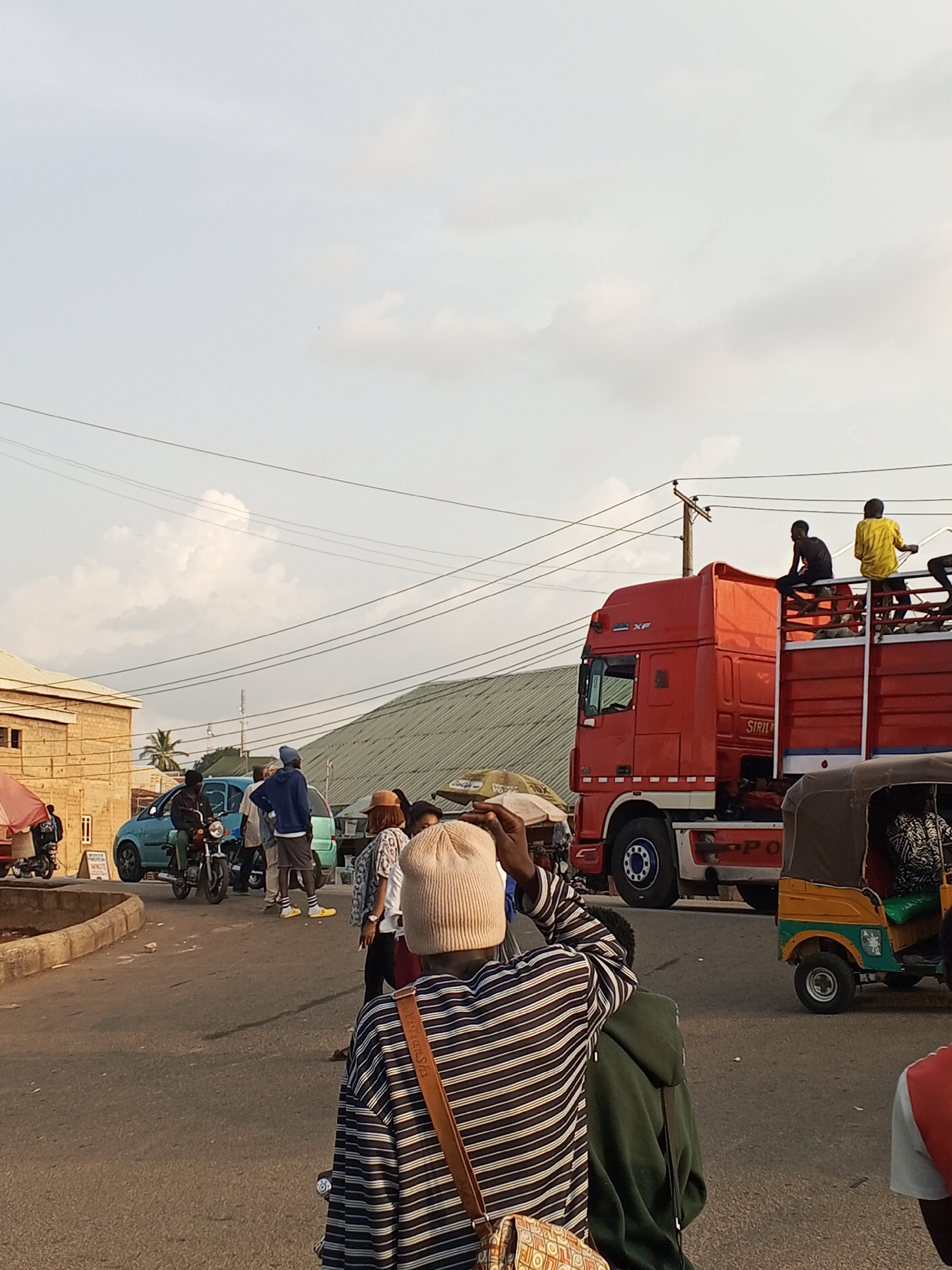 The image shows a busy street scene in Gwong, Nigeria. In the foreground, a person wearing a striped shirt and a beige beanie is facing away from the camera. A red truck is in the mid-ground with two people sitting on top of the back of the truck. A tuk-tuk is to the right of the truck. Other people are standing near cars and motorbikes on the left side of the street. Power lines are visible overhead. Buildings are in the background, including one with a corrugated metal roof. The sky is overcast with clouds.