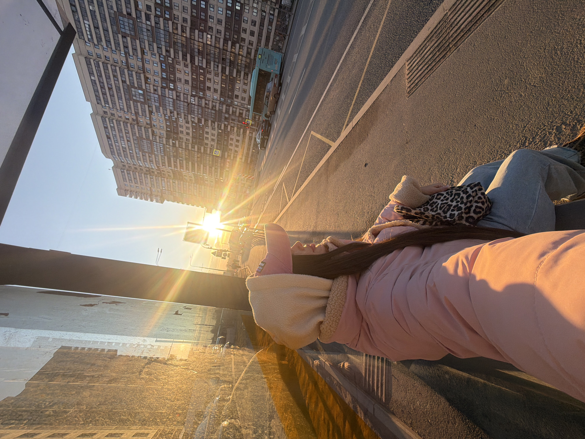 A woman lies on a concrete surface in what appears to be an urban setting, possibly a bridge or elevated walkway. She wears a pink coat with a cream-colored fur-lined hood, light blue jeans, and a pink hat. She is holding a beige and leopard print bag close to her body.
The background shows a modern high-rise apartment building. A road runs parallel to the building. The sun is prominent in the frame, casting a strong glare. Based on the architecture and the style, this may be located in the city of Saint Petersburg, Russia. There is a vehicle on the road. In the foreground, there appears to be a barrier or a reflective surface.