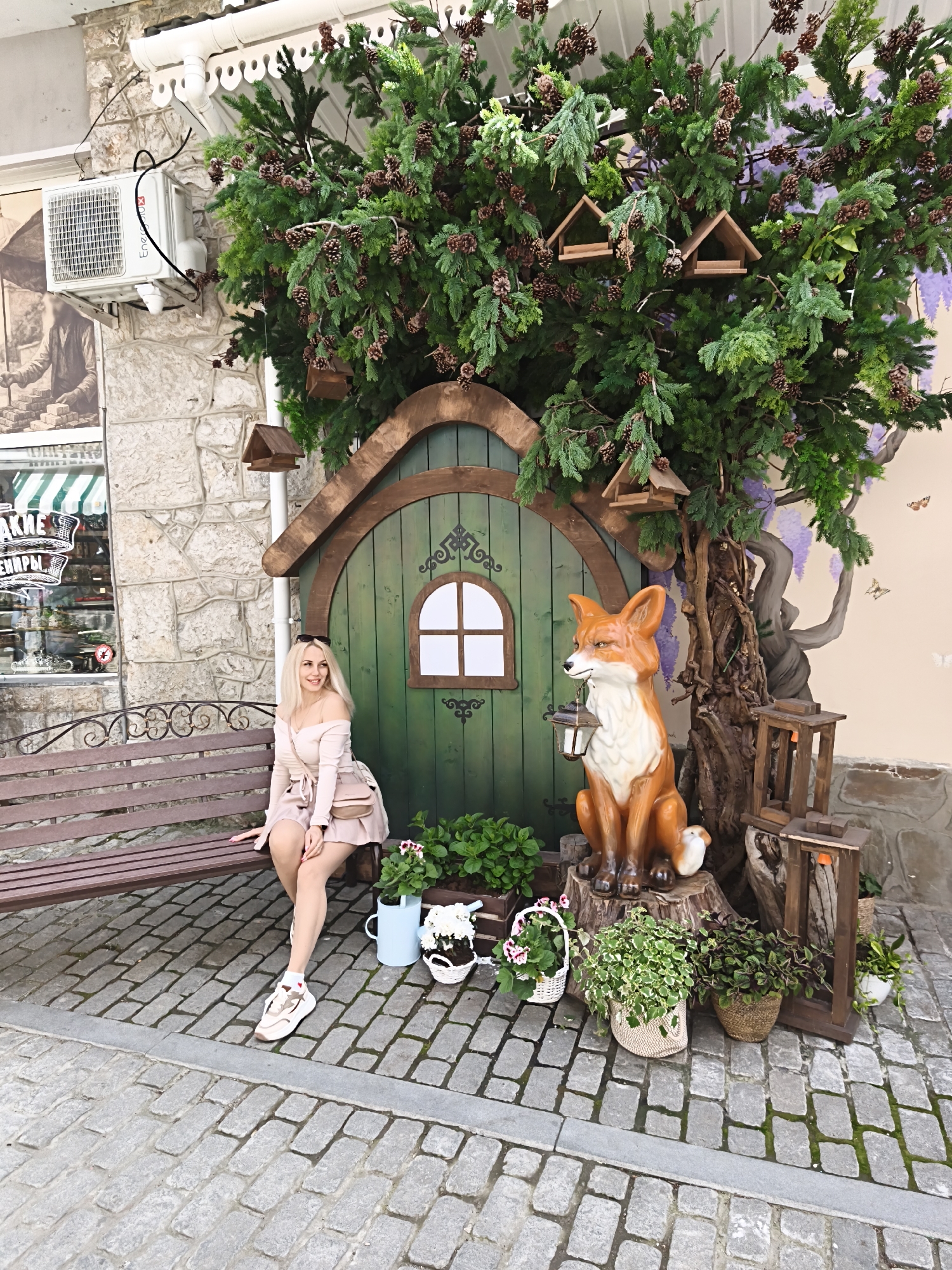 A woman is sitting on a wooden bench next to a whimsical scene in the city of Алупкинский городской совет, Ukraine. The scene features a small, green, arched door resembling an entrance to a fairy-tale house, adorned with a surrounding tree that has birdhouses nestled among its branches. Next to the door stands an orange fox statue holding a lantern. The ground is paved with cobblestones, and there are various potted plants and decorative items around the base of the scene. In the background, a building with a stone facade and an air conditioning unit is visible. The storefront of a shop is also partially visible.