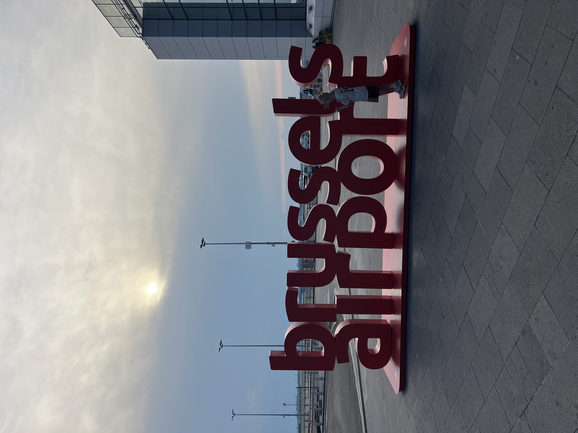 In Zaventem, Belgium, a large, bright red sign spelling out "brussels airport" is prominently displayed on a paved outdoor area. The sign is composed of individual, three-dimensional letters, with "brussels" in the top row and "airport" in the bottom row. A young boy, wearing a white t-shirt with a number and stripes, and black shorts, is standing by the letter "s" in "brussels", appearing to interact with the sign. In the background, a modern, multi-story building with glass windows and a gray facade is visible, alongside streetlights and a road. The sky is cloudy, with the sun peeking through in the upper left quadrant. The scene appears to be set at an airport terminal.