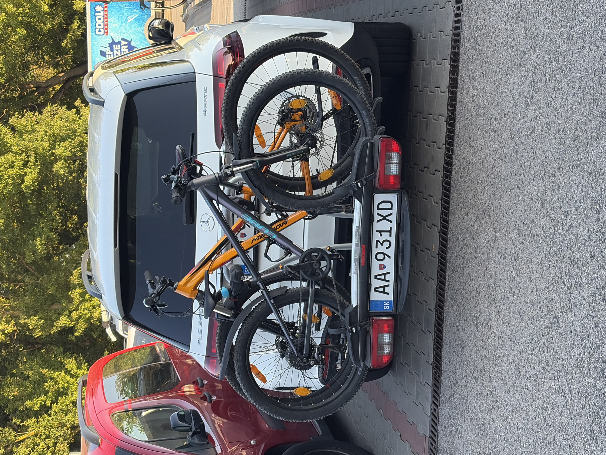A white Mercedes-Benz SUV is parked on a paved area with interlocking stone tiles. The rear of the SUV is visible, along with a portion of a red vehicle to its left. Two bicycles are mounted on a black bike rack attached to the rear of the SUV. The license plate of the SUV reads "AA 931XD" with "SK" on the left side, indicating it's a Slovakian plate. The bicycles are a dark grey mountain bike and an orange mountain bike. The background shows green trees. The scene appears to be in a parking lot or roadside area.