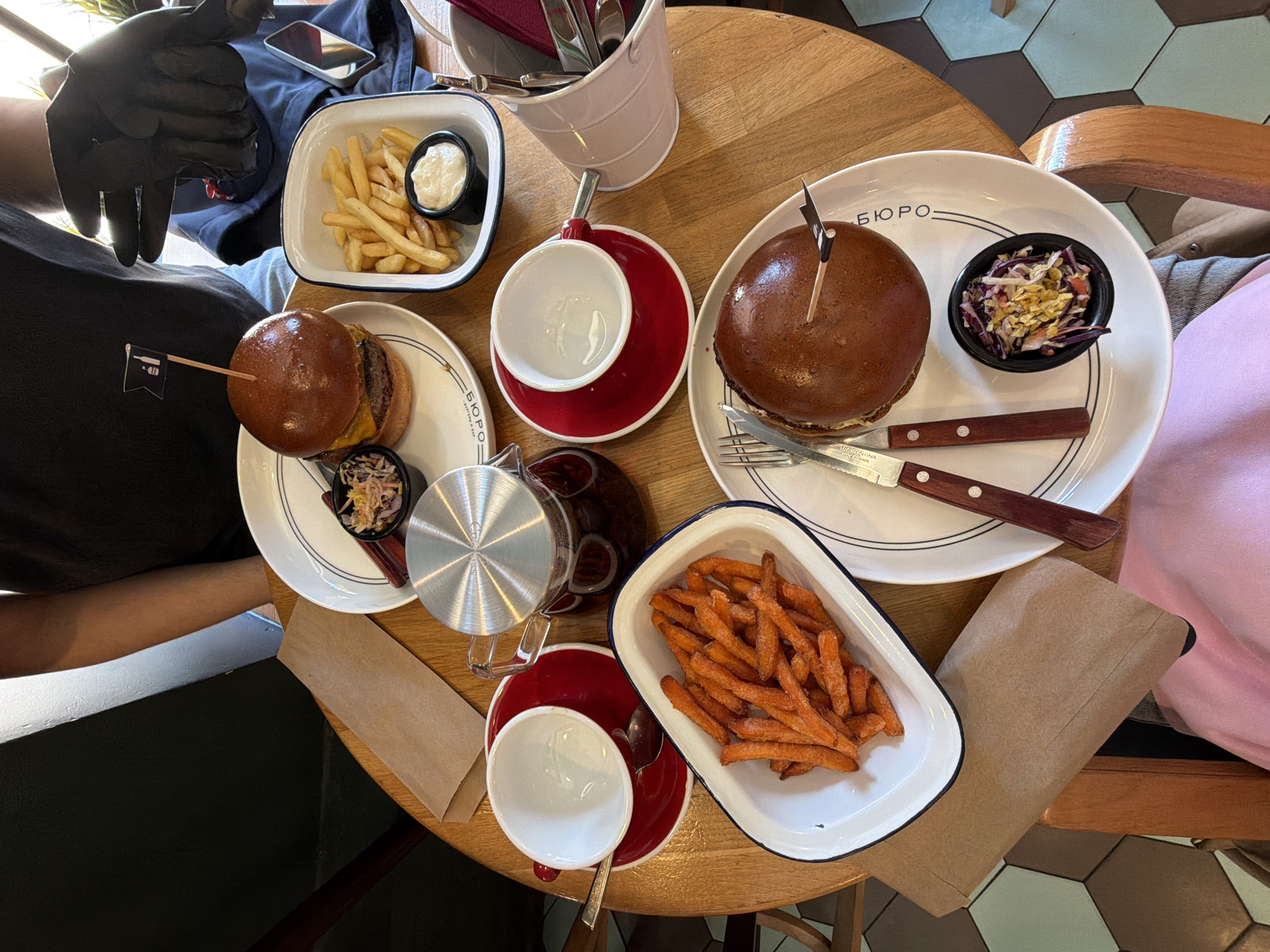 The image shows a table in a restaurant, presumably located in Saint Petersburg, Russia (based on the Cyrillic characters on the plates). The table is round and made of light-colored wood. It is covered with food and dining items.
There are two burgers, each on a white plate with the restaurant's name ("БЮРО") printed on it. One burger has a small side of coleslaw. There are also two plates of french fries, one with regular fries and a dipping sauce, and the other with sweet potato fries. Additionally, there is a tea set with a teapot, two cups and saucers, and a bucket holding cutlery.
Two people are visible at the table, both wearing casual attire. The person on the left is wearing a black t-shirt and black gloves. The person on the right is wearing a light pink sweater.
The table setting suggests a casual dining experience with a focus on burgers and fries. The flooring visible beneath the table consists of dark hexagonal tiles.