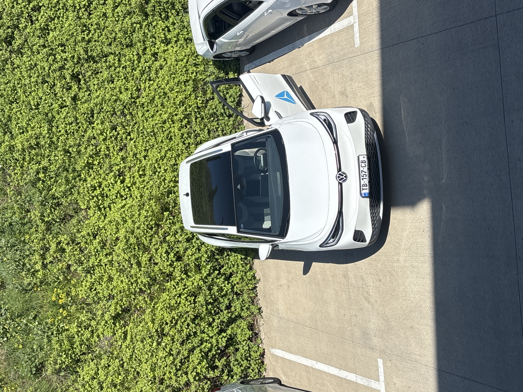 An overhead, high-angle shot captures a white Volkswagen ID.4 electric SUV parked on a concrete surface, adjacent to a dense patch of vibrant green foliage. The driver's side door of the ID.4 is open, revealing the car's interior, including the steering wheel and seats. A prominent blue, triangular logo, consistent with the Wolt delivery service branding, is visible on the inside of the open door. The car also features a panoramic sunroof.
The license plate, clearly legible on the front bumper, reads "TB-157-CB" and includes a small "GE" country code, indicating the vehicle is registered in Tbilisi, Georgia. Another white car is partially visible, parked directly behind the ID.4 on the concrete.
The scene is brightly lit by direct sunlight, casting distinct, sharp shadows across the concrete, suggesting it is midday or early afternoon under clear weather conditions. No people are visible.