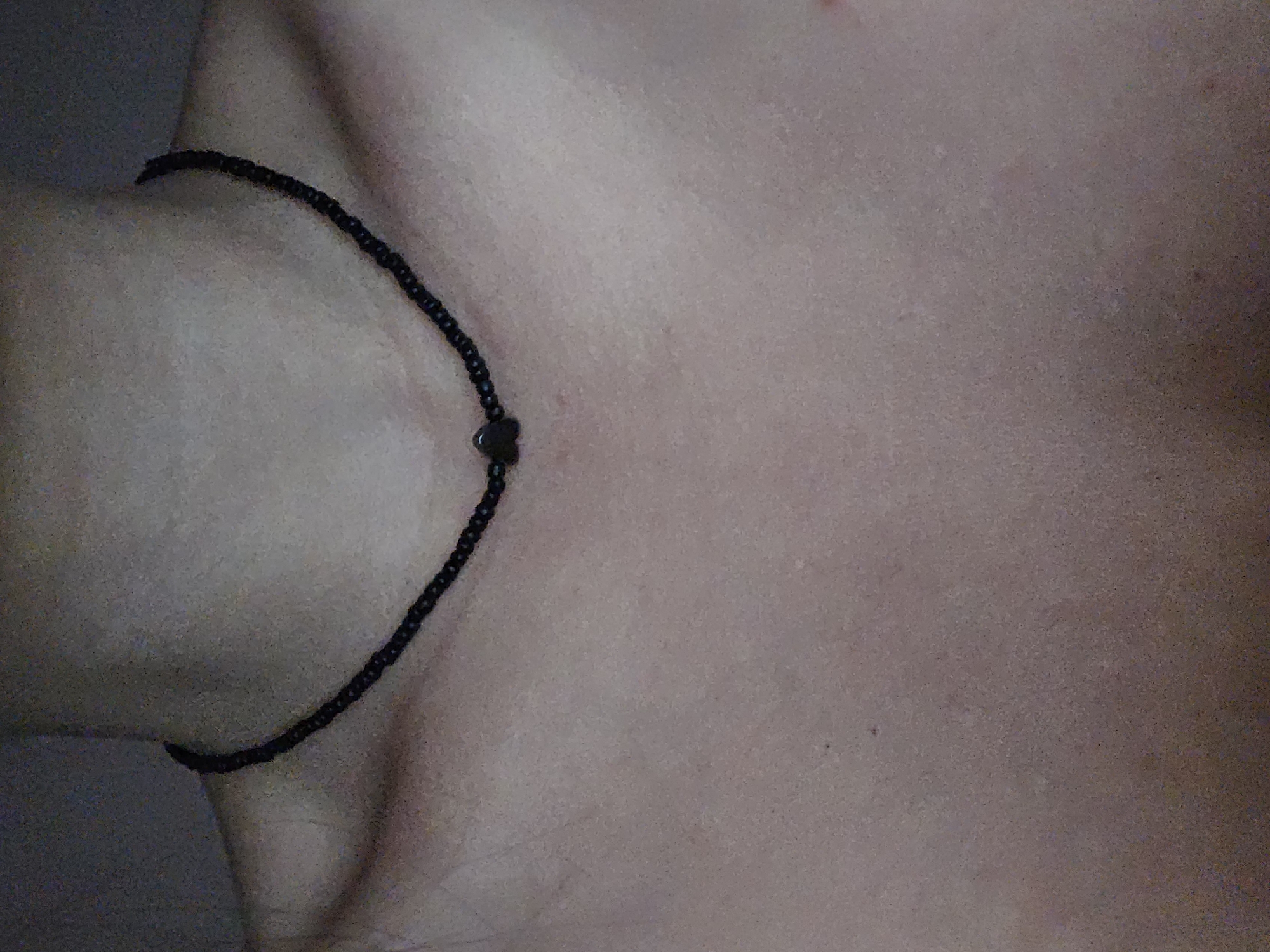 A close-up, dimly lit, and grainy overhead shot focuses on a person's neck and upper chest. A delicate black beaded necklace with a small, dark heart-shaped charm is worn around the neck. The skin texture is visible, with subtle variations in tone and some minor imperfections. The background is dark and out of focus, providing no discernible environmental details. The location of Petropavlivska Borshchahivka, Ukraine, is not visually represented in this image.