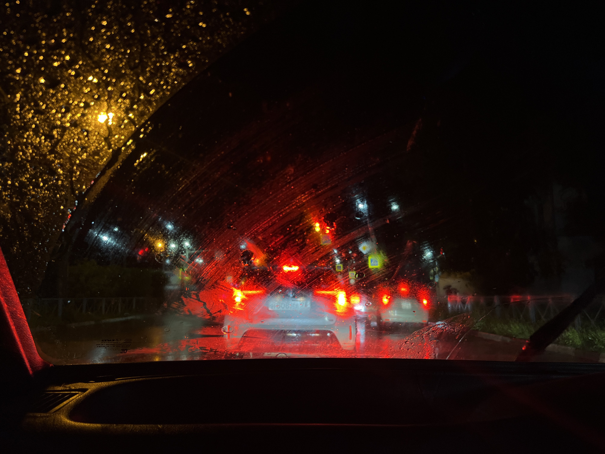 The image depicts a view from inside a vehicle at night, looking through a wet windshield. The windshield is covered in raindrops, which distort the light from street lamps and vehicle lights, creating a bokeh effect. The scene outside shows a road with multiple vehicles, all with their red taillights illuminated, suggesting they are either stopped or moving slowly in traffic. The overall atmosphere is dark and rainy, typical of a nighttime drive in inclement weather. The provided context indicates this scene is located in the city of Belogorsk, Russia, a detail that influences the potential cultural and environmental context of the scene. However, no specific landmarks or identifiers of Belogorsk are visible in the image itself.