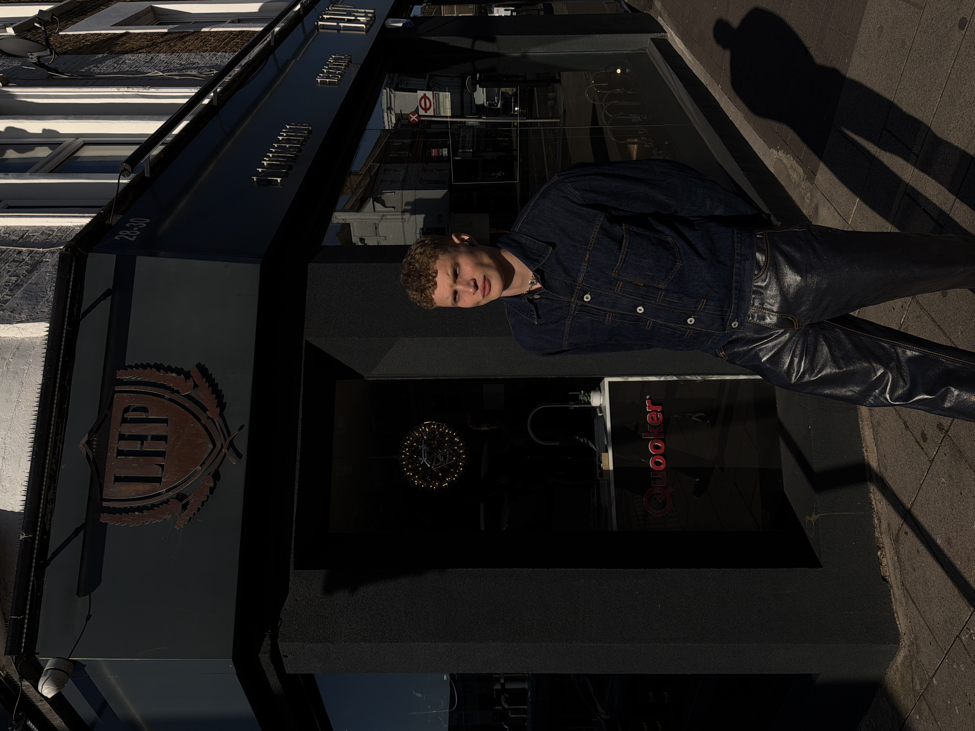An outdoor, full-length shot captures a young man standing in front of a dark, modern shopfront on a sunny day in London, United Kingdom. The man, with short, curly light brown hair, looks directly at the camera with a neutral expression. He wears a dark blue denim jacket with white buttons and shiny, dark leather-like trousers. A subtle necklace is visible around his neck.
Strong sunlight casts a distinct shadow, likely of the photographer, on the paved sidewalk to the right of the man. The building behind him features large display windows that reflect the street and buildings opposite. Visible in the reflections are classical architectural details and a red London Underground roundel sign, confirming the location.
To the left of the man, on the dark facade of the building, a bronze-toned shield-shaped logo featuring the stylized letters "LHP" is mounted. The building number "28-30" is also visible on the darker section above the logo. Inside one of the shop windows, a spherical light fixture with numerous small lights can be seen. Further inside, a red "Quooker" logo is displayed. The overall scene depicts a posed portrait in an urban setting.