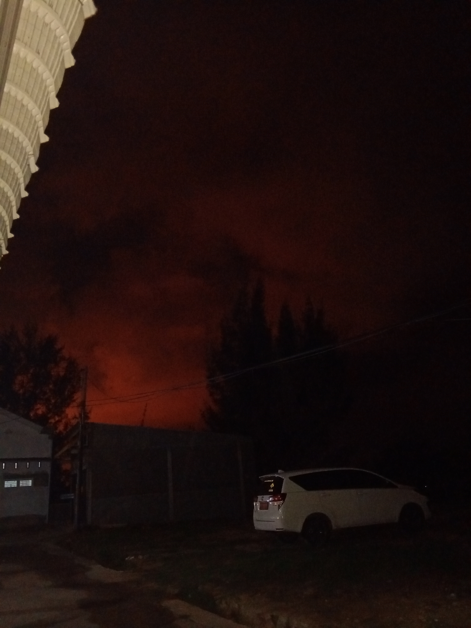 The image shows a nighttime scene in what appears to be a residential area. The sky is a dark reddish-orange, possibly indicating a fire or other atmospheric phenomenon. On the left, there's a structure, possibly a house or building, with visible windows. In the foreground, there is a car parked on what looks like a driveway or a grassy area. Behind the car and the structure, there is a fence or wall, and further back, there are trees silhouetted against the reddish sky. The overall impression is one of an unusual or potentially ominous situation due to the sky's color. While it is difficult to definitively confirm the location as Lhokseumawe, Indonesia without additional contextual information or landmarks, the general characteristics could align with a residential area in that region.