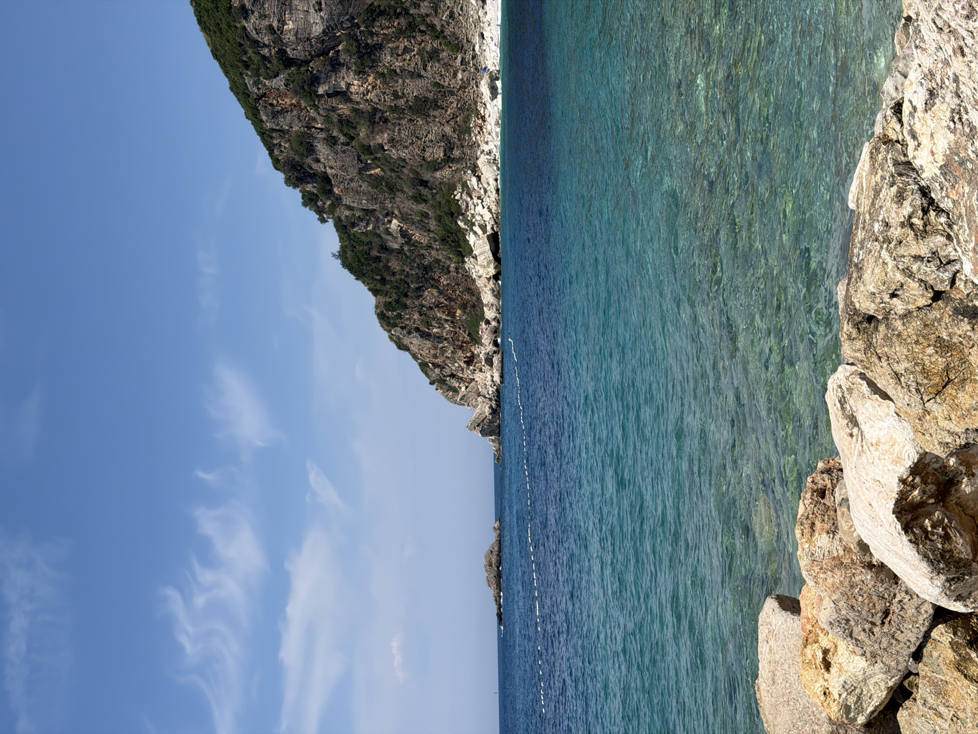 The image captures a scenic view of the Adriatic Sea from the coast of Perazića Do, Montenegro. In the foreground, large, weathered rocks are piled along the shoreline, with the clear, turquoise water gently lapping against them. The sea extends into the midground, transitioning to a deeper blue further offshore. A white buoy line stretches across the water, marking a boundary.
To the left, a small, rocky islet rises from the sea. In the background, a dramatic, verdant cliff face dominates the right side of the frame, its rugged surface covered in dense green vegetation. The sky above is a bright blue, with wispy white clouds scattered across it. A single person, visible as a small blue dot on the rocky shore near the base of the cliff, adds a sense of scale to the vast landscape. The overall impression is one of a serene and natural coastal environment.