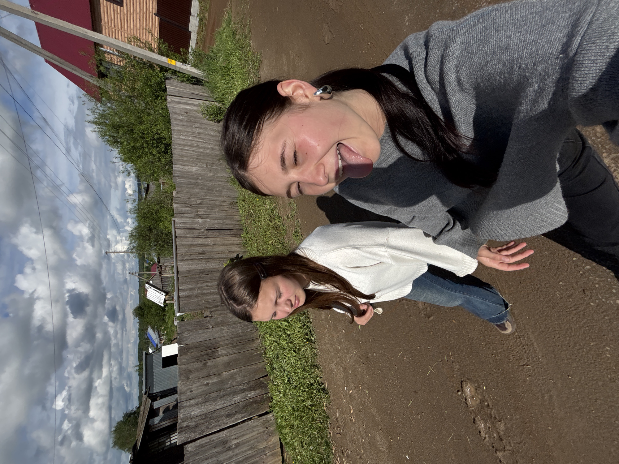 Two young women are captured outdoors in what appears to be a rural or semi-rural setting. The woman in the foreground, closer to the camera, has dark hair and is wearing a grey long-sleeved sweater and dark pants. She is making a playful face, sticking out her tongue and closing her eyes, with a large, silver-toned ear cuff visible on her ear. The second woman stands slightly behind and to the left of the first, wearing a white long-sleeved sweater, blue jeans, and light-colored sneakers. She has brown hair and is looking downwards or to the side, with a neutral expression.
They are standing on a dirt path or unpaved road. To their left, a weathered wooden fence runs alongside the path, bordered by green bushes and vegetation. In the background, power lines stretch across the frame, supported by wooden utility poles. Beyond the vegetation and fence, distant houses, some with red roofs, are visible under a partly cloudy sky. The bright daylight is evident from the shadows cast on the ground and subjects, suggesting it is midday or early afternoon. The weather is clear but with significant cloud cover. A small yellow sticker with unreadable text is present on one of the wooden poles. The scene's elements, including the wooden fence, dirt road, and type of housing, are consistent with a village environment in Foki, Russia.