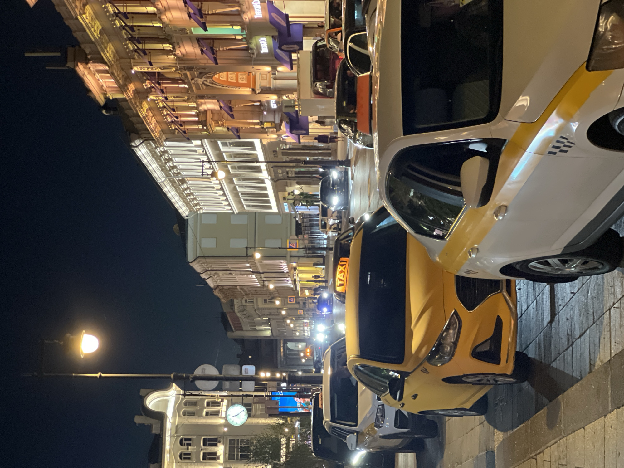 The image depicts a nighttime street scene in Moscow, Russia, specifically identified as Stoleshnikov Lane. The perspective is looking down the lane, likely from the area near Petrovka Street towards Bolshaya Dmitrovka.
In the foreground, a white sedan taxi, bearing a yellow stripe and a black checkered pattern on its side, is partially visible on the right. Immediately to its left and slightly forward is a yellow sedan taxi, facing towards the viewer. The car's front grille and headlights are prominent.
The midground features a line of stationary or slow-moving vehicles, predominantly taxis, extending into the distance. These include additional yellow sedans, a white SUV with visible headlights, and other vehicles. Several taxis display illuminated "TAXI" signs on their roofs. Headlights from oncoming vehicles are visible further down the street.
On the left side of the street, a multi-story white building with ornate architectural details stands illuminated. A large, circular clock face on this building shows the time as approximately 11:20 or 11:25. A streetlamp with a single glowing bulb is positioned in the immediate foreground, casting light onto the paving stones of the sidewalk.
On the right side of the street, a series of multi-story buildings display intricately detailed facades, lit windows, and purple awnings over ground-level establishments. Visible store signage includes "Tiffany & Co." and "ETRO."
The street surface is paved, and sidewalks with patterned paving stones are visible on both sides. Multiple pedestrians are discernible on the sidewalks in the background. The sky is dark blue, characteristic of nighttime. The entire scene is brightly illuminated by a combination of streetlights, building facade lighting (varying from warm yellow to cool white), and vehicle lights. Traffic signs and other urban fixtures are present throughout the scene.