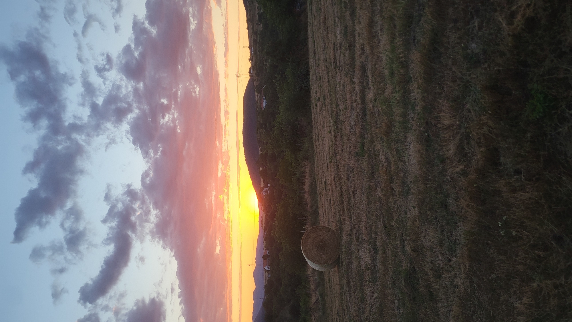 The image depicts a rural landscape at sunset. A single, round bale of hay sits in the foreground on a dry, grassy field. In the mid-ground, a line of trees separates the field from a gentle, rolling hill dotted with scattered houses and structures. A prominent tower, likely a cell tower or electrical pylon, is visible on the hilltop. The background is dominated by a dramatic sky filled with clouds illuminated by the setting sun, casting hues of orange, yellow, and pink. The overall scene suggests an agricultural area in the vicinity of Rechitsa, Bulgaria, given the mention of the city. There are no people or animals visible in the frame, and the environment appears peaceful and serene.