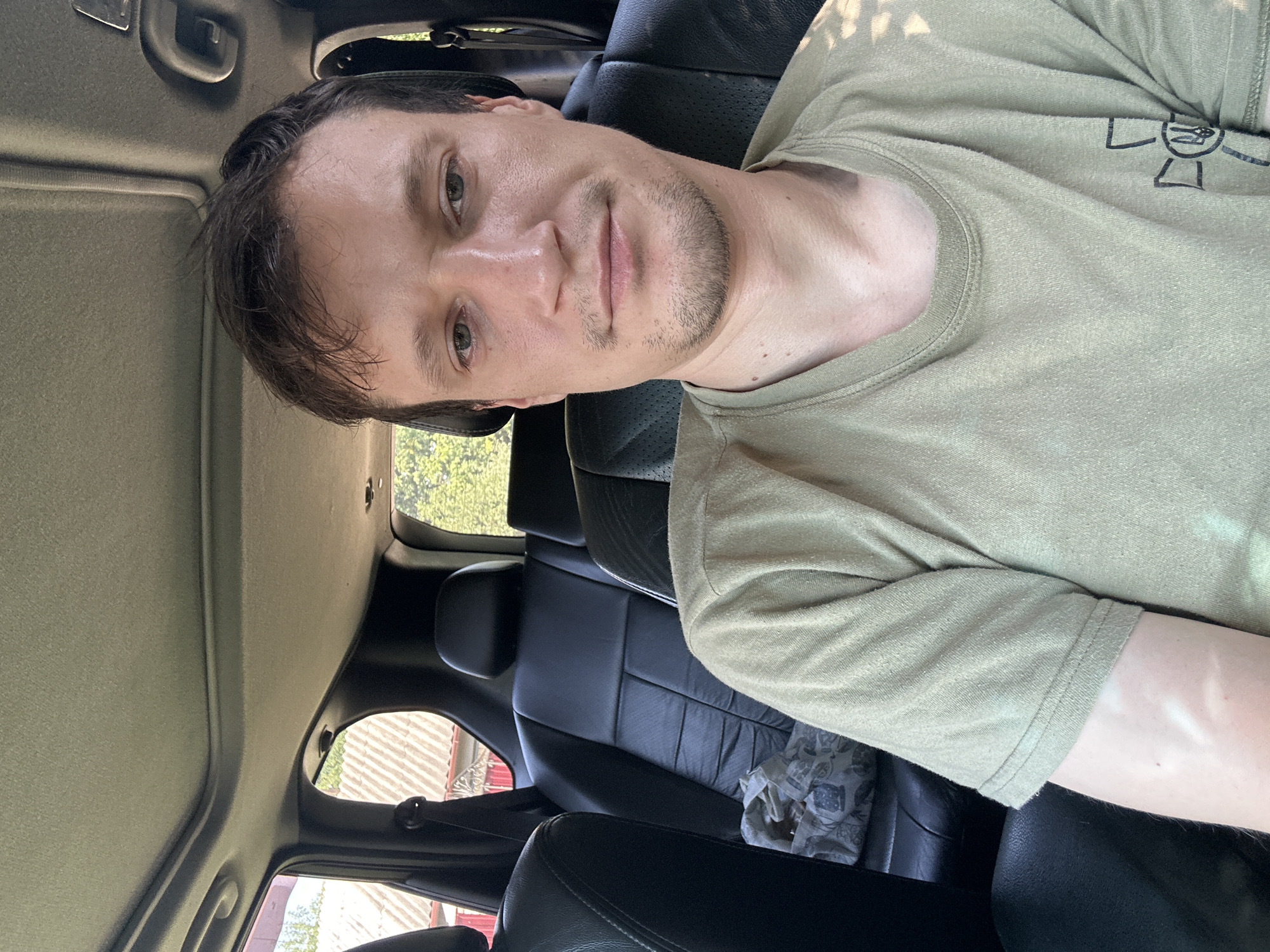 A medium shot shows a man in his late 20s to early 30s sitting in the back seat of a car. He has short brown hair, light skin, and stubble on his chin. He is wearing a light green t-shirt with a black emblem on the left side of his chest, which appears to be a Ukrainian trident within a cross. The interior of the car is visible, with black leather seats and a sunroof overhead. Outside the car, through the windows, trees and a building with a tiled roof can be seen, suggesting a rural or suburban setting, possibly in Shatava, Ukraine. The lighting indicates it is daytime. The man is looking directly at the camera with a neutral expression.