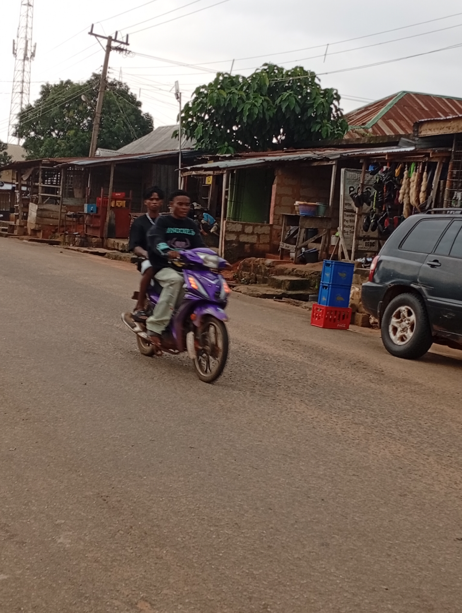 The image depicts a street scene in Nsugbe, Nigeria. In the foreground, two young men are riding a purple motorcycle. The rider in front is wearing a black t-shirt, and the passenger behind him is wearing a black long-sleeved shirt and light-colored pants. They are moving on a paved road. To the right of the road, a dark-colored SUV is parked. Behind the motorcycle and SUV, there are several stalls or small shops with corrugated metal roofs. One stall has various items hanging for display. Red and blue plastic crates are stacked on the ground near the parked SUV. In the background, trees and a tall telecommunications tower are visible. The sky is overcast.