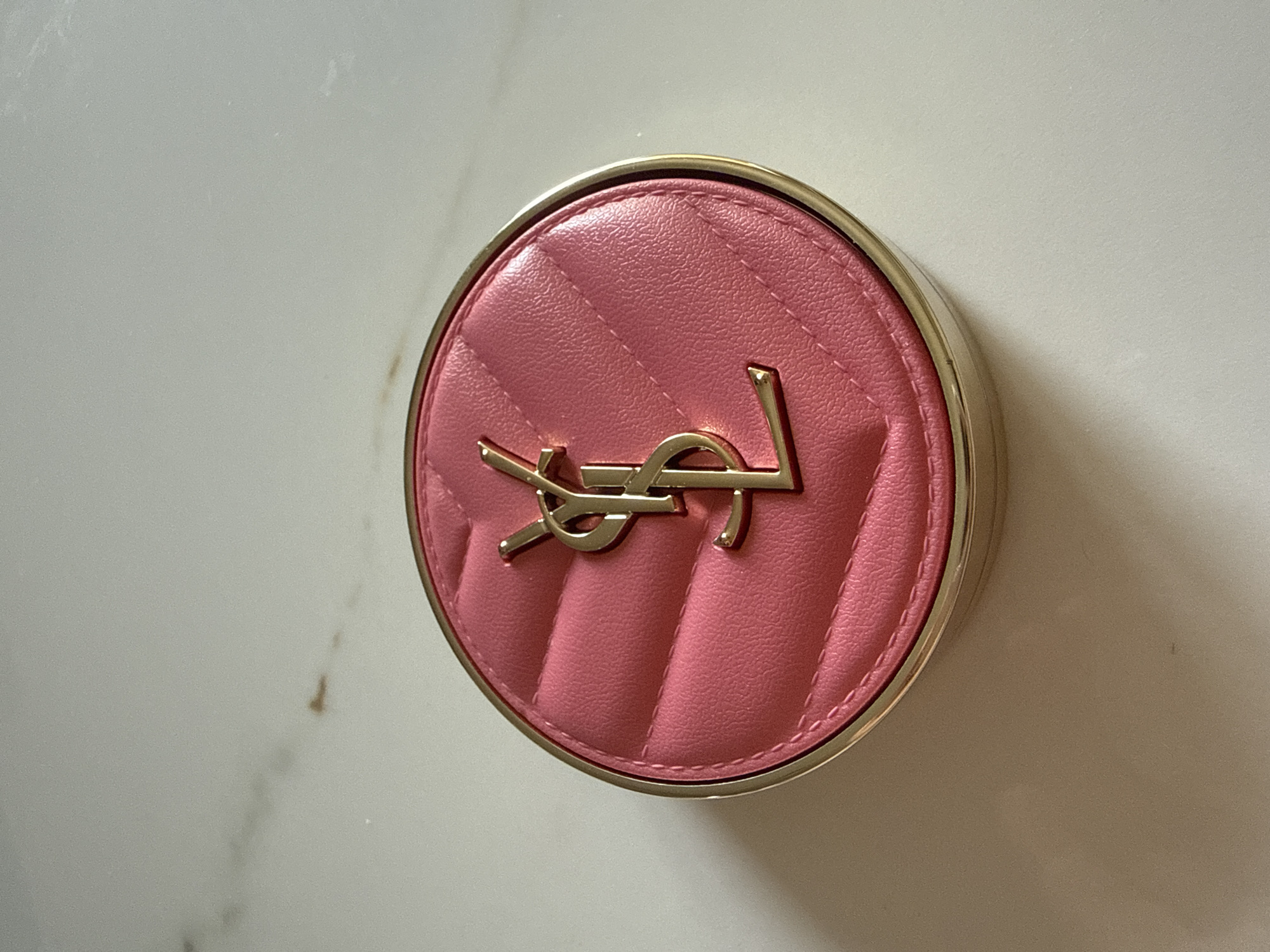 The image shows a YSL (Yves Saint Laurent) compact on a surface. The compact is round with a gold-colored rim and a pink quilted top featuring the YSL logo in gold. The surface appears to be white with subtle marble-like patterns. The setting is indoors.