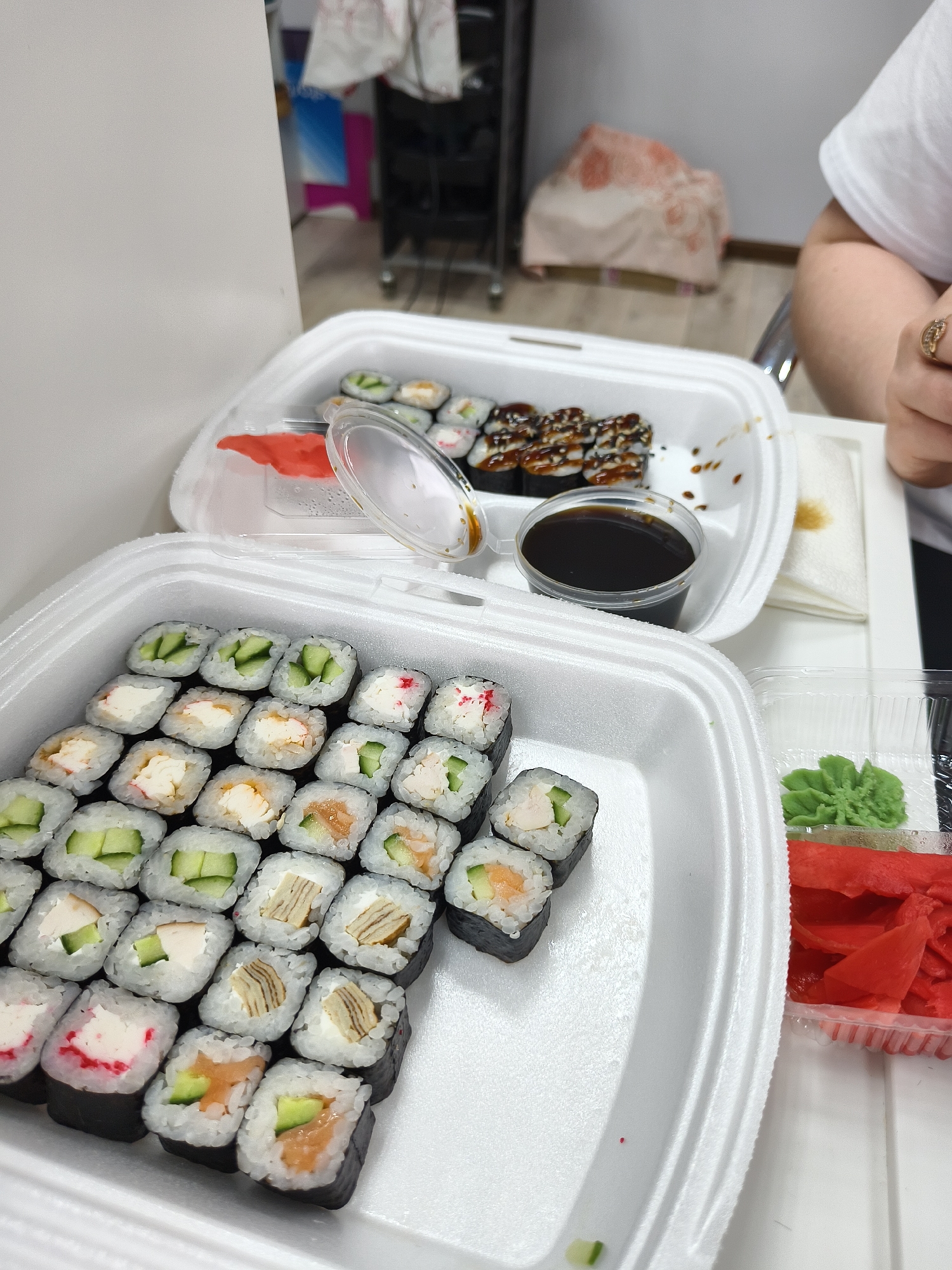 The image displays a close-up, overhead view of two white Styrofoam take-out containers filled with an assortment of sushi rolls, placed on a white surface, likely a table.
In the foreground, a large container is packed with numerous maki sushi rolls. These rolls exhibit various fillings, including cucumber, cream cheese often paired with crab stick, salmon, and what appears to be tamago (Japanese omelet). The rolls are neatly arranged in rows, showcasing the different ingredients.
Behind the large foreground container, a second, partially opened Styrofoam box contains fewer sushi rolls, notably some topped with a dark, glistening sauce, indicative of eel sushi. This container also holds a small plastic cup filled with dark soy sauce, with its clear lid partly open.
To the right of the main sushi box, a smaller clear plastic container holds a dollop of bright green wasabi and a generous portion of vibrant red pickled ginger. A white folded napkin, soiled with a brown stain, rests next to the upper sushi box.
A person's right arm and hand, wearing a ring and part of a white t-shirt, are visible on the far right edge of the frame, suggesting someone is present and potentially about to enjoy the meal. The background is blurred, showing a generic indoor setting with a white wall, a dark wheeled cart, and a light-colored bag. No discernable text is visible in the image.