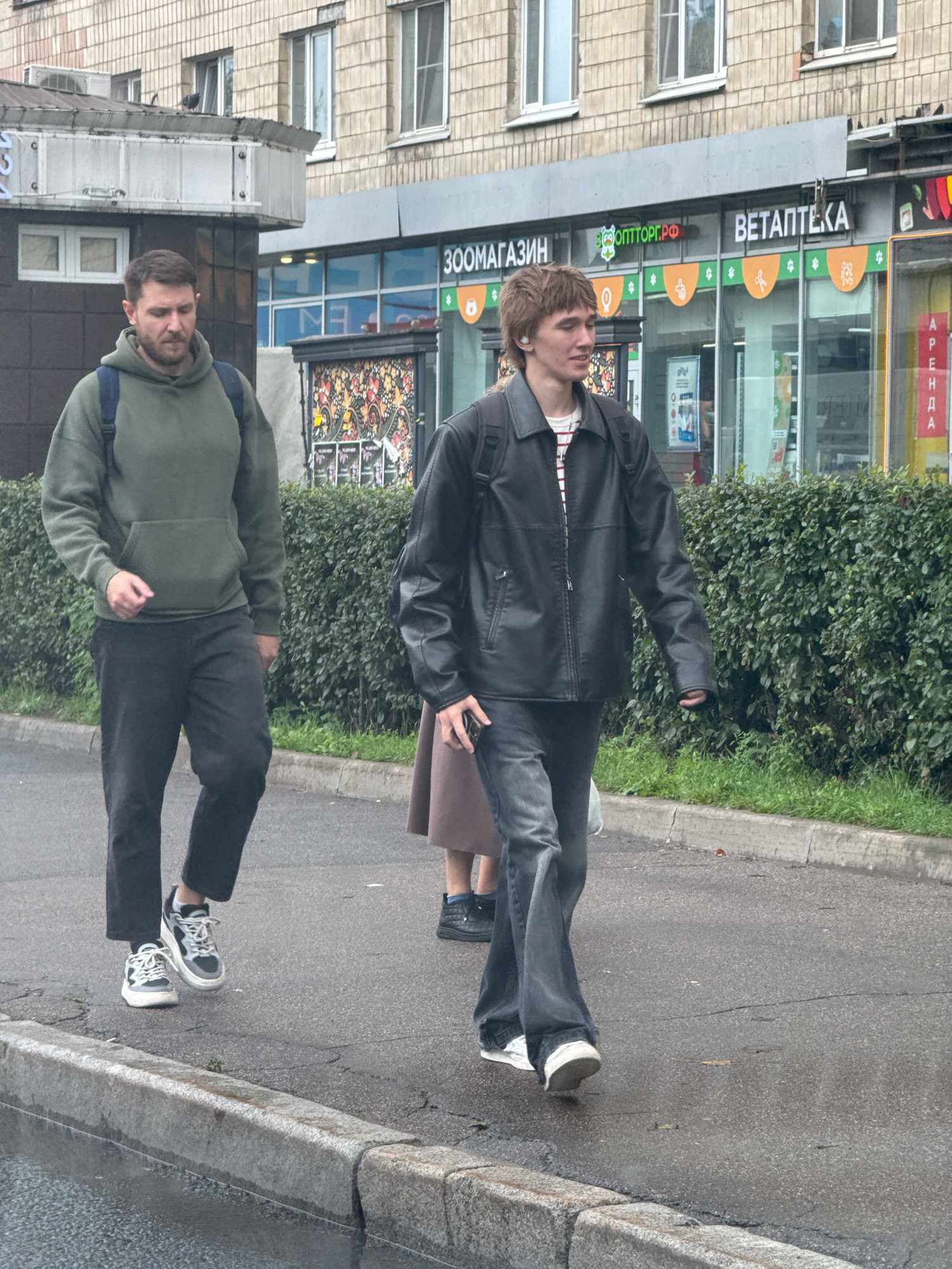 Two men are walking on a wet sidewalk in front of shops in Saint Petersburg, Russia. The man on the left wears a green hooded sweatshirt, black pants, and grey and white sneakers. He has a blue backpack. The man on the right wears a black leather jacket over a striped shirt, dark flared pants, and white sneakers. He also has a backpack. A woman in a brown skirt is partially visible between them. The shops have signs in Russian, including "ЗОOPTTOPГ.РФ" and "ВЕТАПTEKA". A dense green hedge runs along the sidewalk behind the people. The sky is overcast.