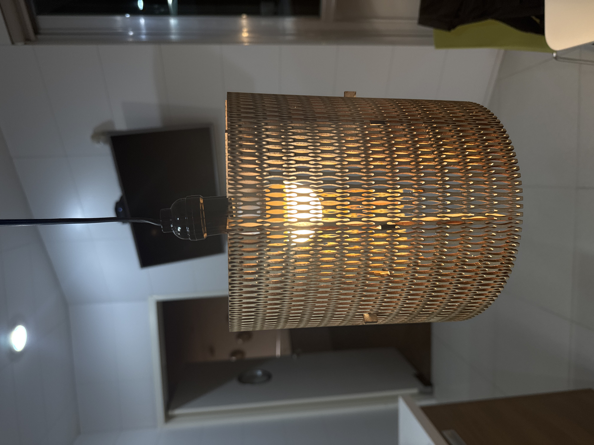 The image, captured in San Bernardino Tlaxcalancingo, Mexico, showcases an illuminated pendant light fixture indoors. The primary subject is a cylindrical lampshade crafted from a light brown material, likely wood or thick cardboard, featuring an intricate laser-cut or perforated pattern. This pattern consists of numerous rows of small, elongated, interlocking shapes, creating a textured, woven appearance. An incandescent bulb glows brightly within the shade, casting a warm, golden light that highlights the detailed pattern and projects through its openings. The bulb is housed in a simple black socket, from which a black electrical cord extends upwards.
The background reveals a modern indoor setting. To the left of the light fixture, a light-colored wall displays a dark, rectangular object, possibly a wall-mounted flat-screen television or monitor. A small, circular, intensely bright light source is also visible on this wall. To the right, indistinct elements suggest further parts of the room, such as cabinetry or an open doorway. The ambient lighting indicates that it is likely evening or night, with artificial light sources providing the primary illumination. No specific activity or event is taking place, and no visible text is present. The scene conveys a clean, contemporary aesthetic.