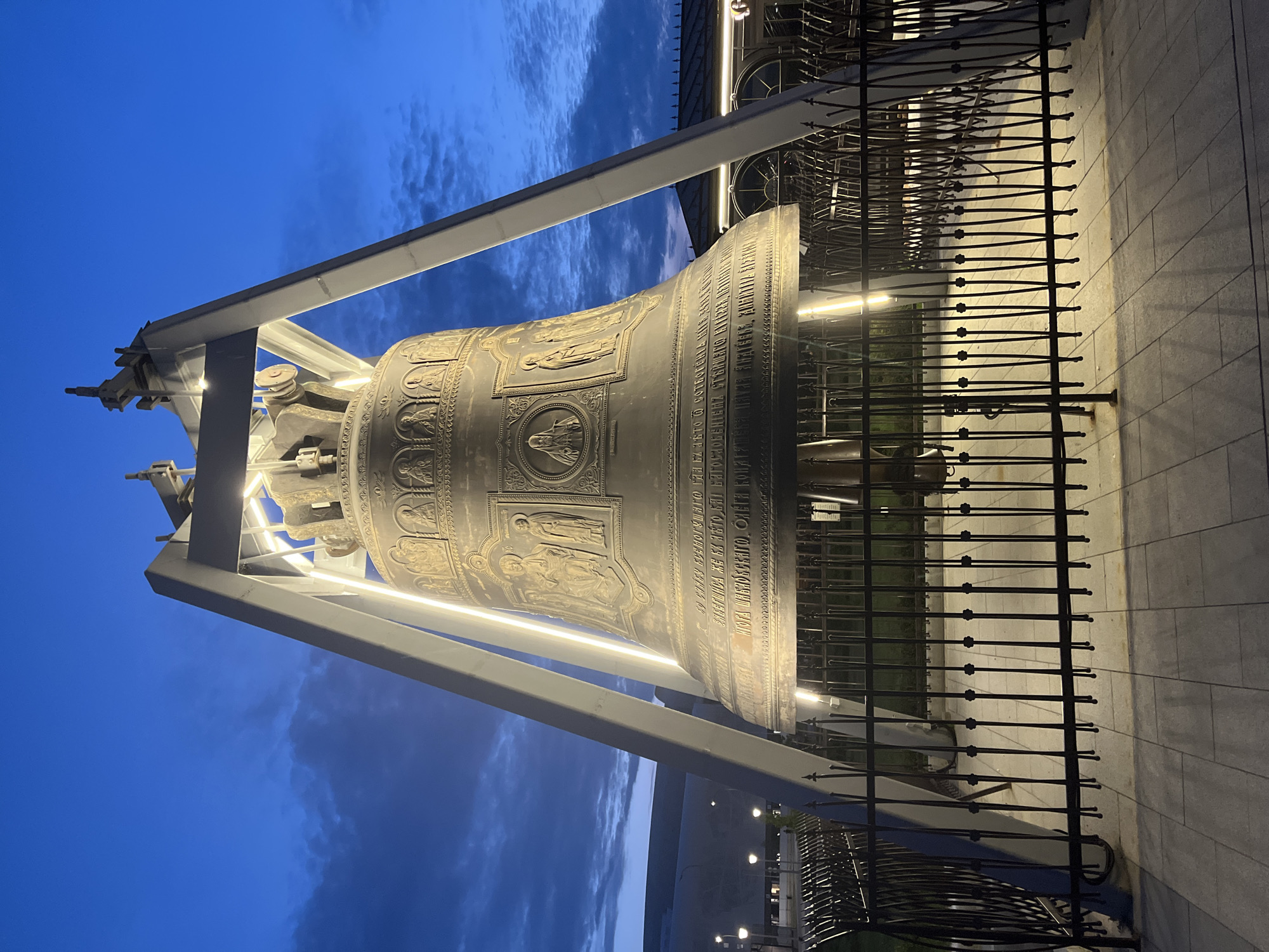 This image shows a massive, ornate bronze bell suspended within a metal frame against a twilight sky. The bell is adorned with intricate relief carvings depicting religious figures and scenes, and also features text in Cyrillic script around its circumference. The sky is a deep blue with scattered clouds, suggesting dusk or early evening.
The bell is surrounded by a black metal fence with decorative elements, and a paved walkway is visible on the right side of the frame. Lights are illuminating the bell and its surroundings, casting long shadows on the pavement. The overall scene evokes a sense of grandeur and historical significance, possibly in a religious or cultural landmark setting.
The visible text appears to be in Russian and likely contains inscriptions related to the bell's history, dedication, or religious imagery. While no people are visible, the presence of a large, imposing object like this bell suggests a public space, possibly a square or the grounds of a cathedral or museum in Nizhny Novgorod, Russia. The time of day and the lighting contribute to a dramatic and somewhat solemn atmosphere.