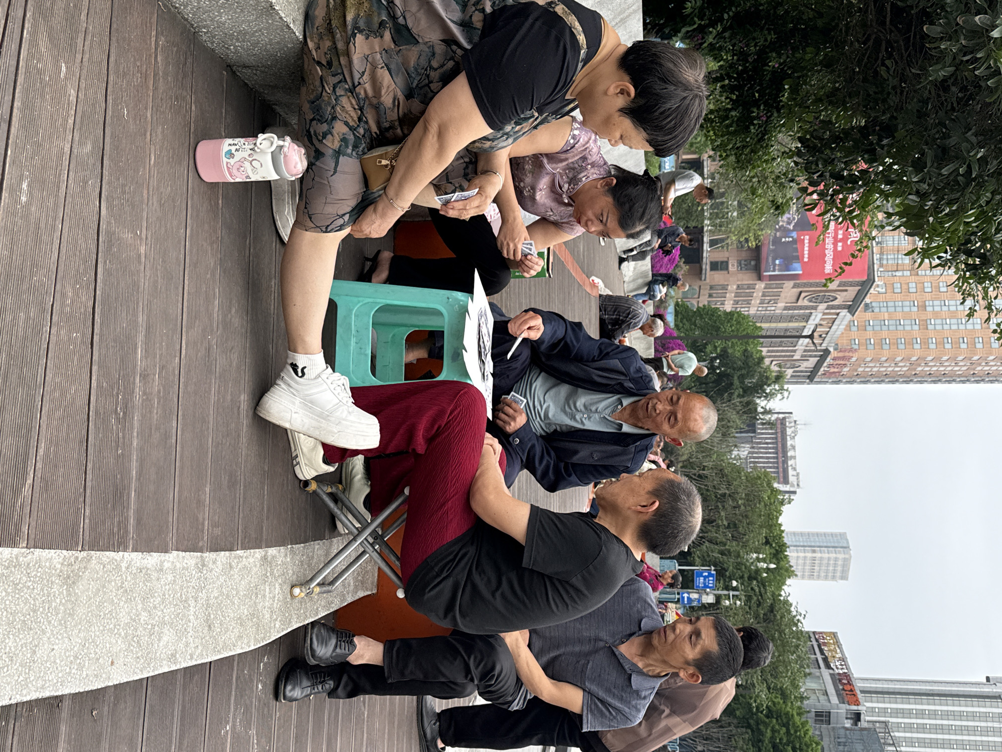 A group of people are gathered on a wooden boardwalk in what appears to be a public space. Several people are seated, with at least two women playing cards on a small green stool. An elderly man in a dark blue jacket and grey undershirt is seated and appears to be engaged in the card game or conversation. Another person is seated in a folding chair, while others are standing or walking in the background. The backdrop includes trees, buildings, and a red billboard with Chinese characters. The image likely originates from the Nan'an District in China, given the context provided.