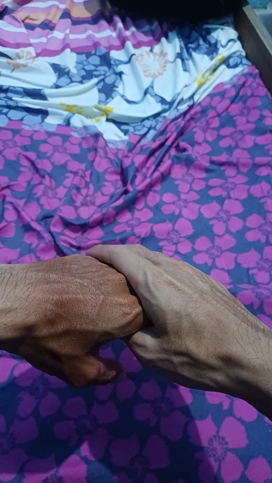 Two hands are clasped together, with one hand appearing darker skinned than the other. The hands are resting on a bed covered with a floral patterned duvet. The duvet has a base color of dark blue with large purple and pink floral designs. The top portion of the bed has a different pattern, featuring stripes of white, pink, and orange, interspersed with blue and light brown floral motifs. The lighting suggests it may be indoors, possibly in a bedroom in Kowangan, Indonesia, given the context.