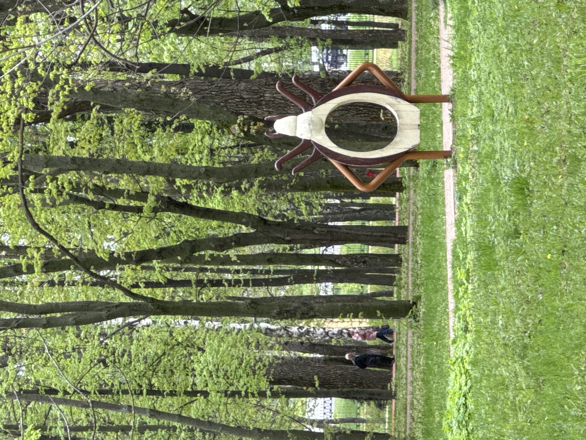 The image shows a park in Nizhny Novgorod, Russia. The focus is on a large sculpture depicting a stylized insect or beetle, constructed from metal and wood. The sculpture has a central circular opening and is mounted on two rectangular supports. The park setting consists of a grassy area, a pedestrian path, and a line of trees with fresh green leaves. In the background, people are seen walking along the path. A metal fence is visible in the distance. The scene is taken in daylight, with natural light illuminating the park.