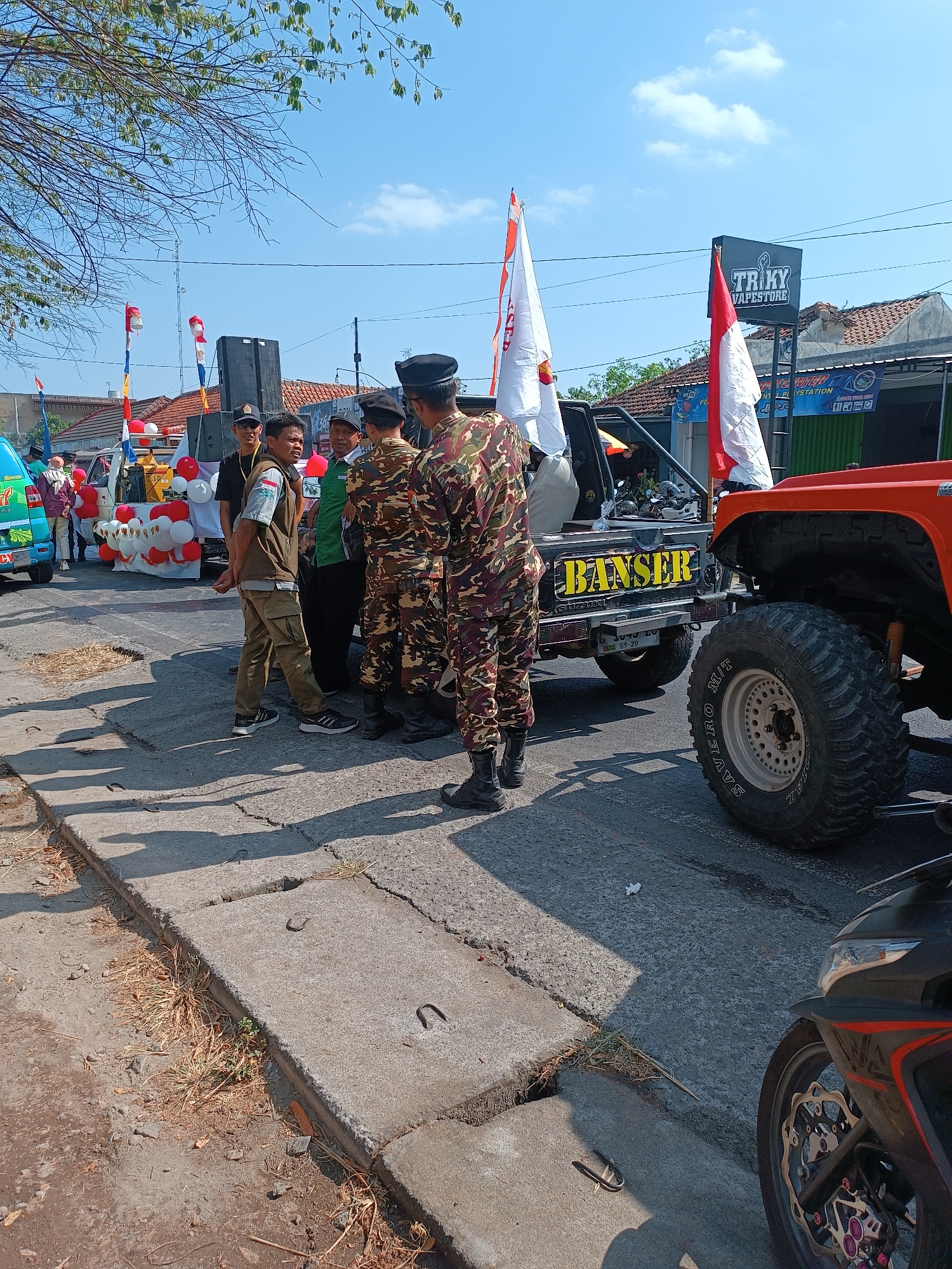 In Imogiri, Indonesia, a group of men in uniform and civilian attire stand on a paved street under a bright, partly cloudy sky. Two vehicles are prominent: a black Suzuki Jimny with "BANSER" in yellow lettering on its side, and an orange off-road vehicle with large tires. The Jimny is decorated with red and white balloons and two Indonesian flags. Several individuals are gathered around the Jimny, some in military-style camouflage uniforms, others in vests or civilian clothes. One man in a vest faces the camera, looking to his left. A large speaker is visible behind the group, and a building with a sign that reads "TRIKY VAPESTORE" is in the background. The street is lined with concrete structures and sparse vegetation. A motorcycle is partially visible in the foreground on the right. The scene suggests an outdoor event or procession.