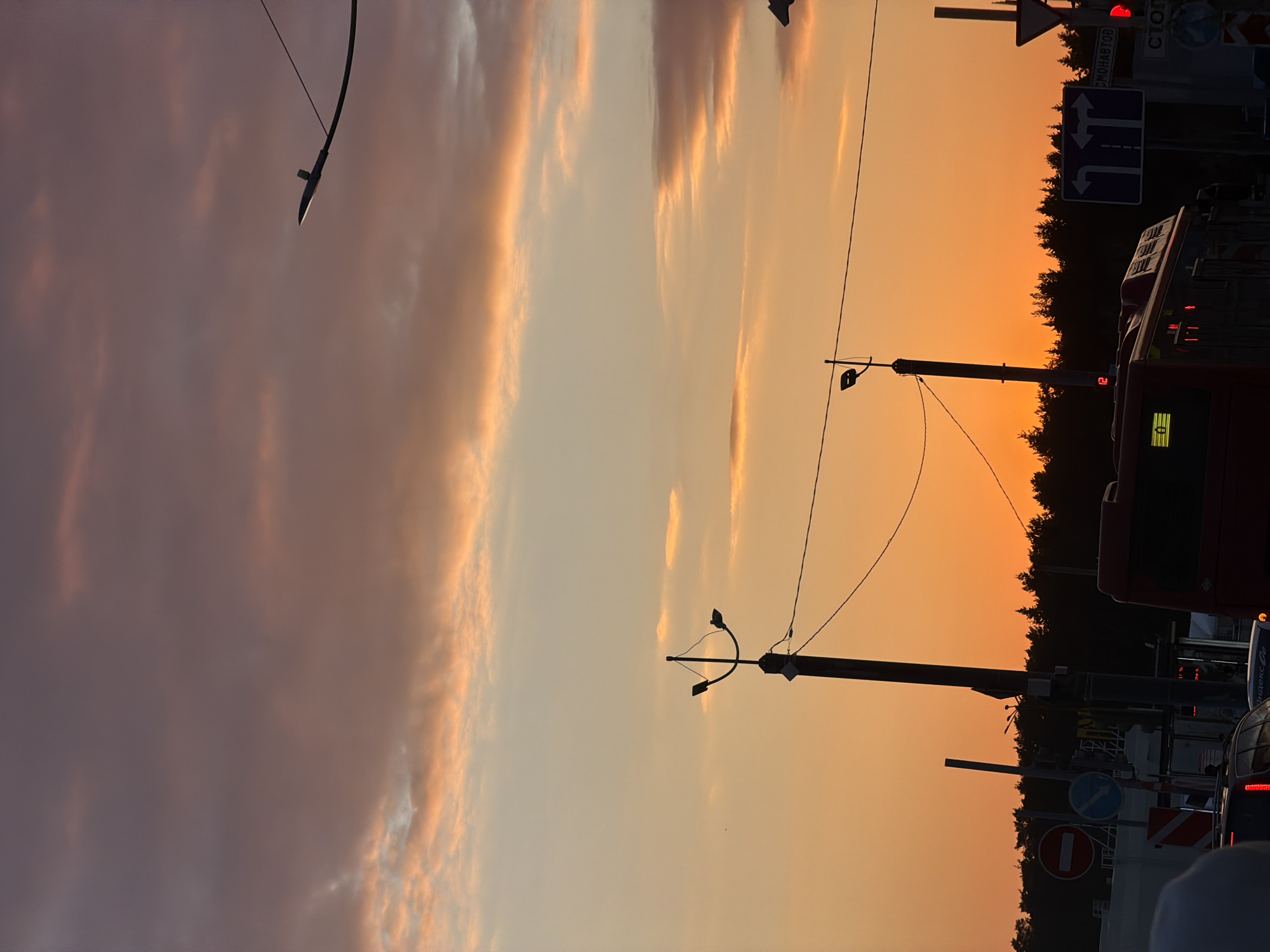 The image depicts an urban street scene at dusk or sunset in Perm, Russia, characterized by a dramatic sky and silhouetted city elements. The dominant feature is the sky, which transitions from bright, warm oranges and yellows along the horizon to softer purplish-gray clouds with golden edges higher in the frame, indicating the golden hour.
Various urban subjects are silhouetted against this colorful backdrop. Several dark utility poles and streetlights, some featuring multiple lamps, are positioned across the scene, connected by overhead wires. To the right, a large vehicle, identifiable as a bus, is partially visible with its dark profile. It displays an illuminated yellow route number '2' and a 'Яндекс Go' (Yandex Go) logo on its side. Other parts of vehicles, including what appears to be the rear lights of a car, are visible at the bottom right.
A dense line of dark coniferous trees forms the horizon line, separating the vibrant sky from the lower, unseen ground level. Several traffic and road signs are visible: a blue rectangular sign with white arrows indicating "straight," "left turn," and "right turn"; a red circular "No Entry" sign; and a red circular "No Stopping" or "No Parking" sign. A red traffic light is also prominent on one of the poles. Visible Cyrillic text includes "СТОП" (STOP) on a red sign, and partially obscured text that appears to read "СМОЛАВТОD" on a white sign. The scene conveys the atmosphere of an active city street as daylight fades.
