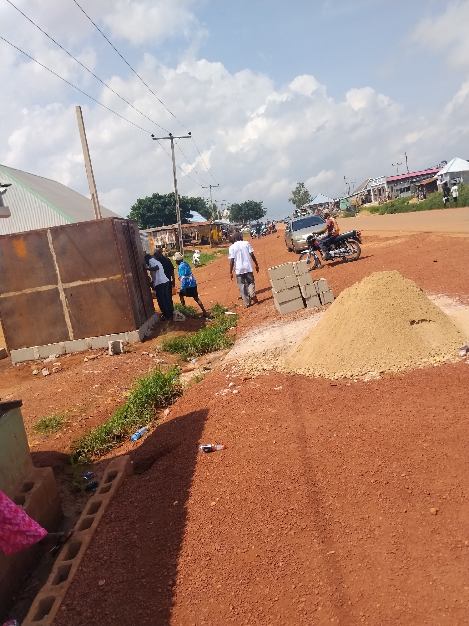 In Durumi, Nigeria, an outdoor scene unfolds under a sky dotted with cumulus clouds. A group of men are gathered near a large, rusted metal structure, possibly part of a construction site. Three men are actively engaged with the structure, one in a white shirt, another in a dark shirt, and a third in a blue and white patterned shirt. To the right, a pile of sand sits near stacked concrete blocks. A motorcycle, ridden by a man in an orange shirt, is parked nearby, with a silver sedan in the background. Several other motorcycles and a few scattered individuals populate the dusty, reddish-brown landscape, which appears to be a road or a construction area. In the distance, various buildings and structures line the roadside, some with corrugated metal roofs. A lone wooden pole with electrical wires stretches across the frame, indicating the presence of utilities. Greenery, including grass and trees, offers patches of contrast to the predominantly earthy tones of the scene. Two plastic water bottles are visible on the ground.