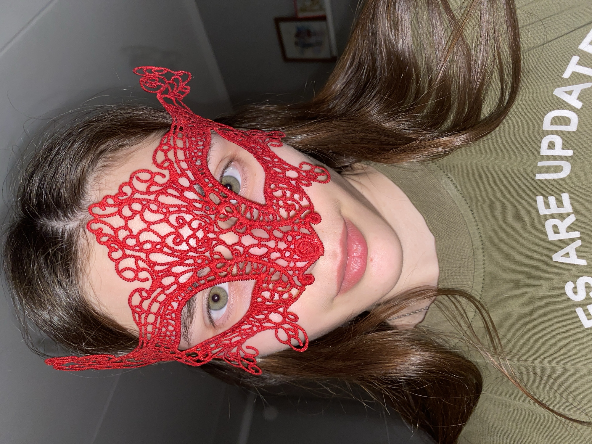 An indoor close-up photograph depicts a person, likely female, from Votkinsk, Russia, posing with their head tilted almost horizontally to the left. The subject has long, brown hair and light green eyes, one of which is visible, looking towards the camera. She wears a striking red lace masquerade mask that covers her forehead and eyes, featuring intricate floral or swirling patterns. Her lips are slightly parted. She is dressed in an olive-green t-shirt, and partially visible white text on the shirt reads "ES ARE UPDAT". A silver chain or necklace can be seen around her neck. The background is a plain, dark gray wall, with a small, indistinct framed picture hanging high up on the wall, partially visible in the upper right. The lighting suggests an indoor setting, possibly with artificial light, creating some soft shadows. The overall impression is one of mystery or playfulness due to the mask.