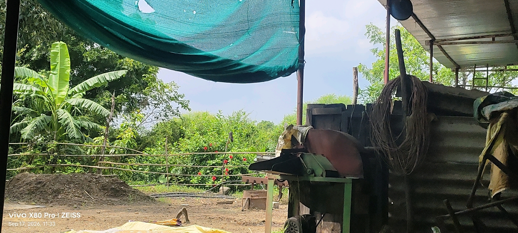 The image depicts an outdoor scene at what appears to be a farm or agricultural area in Guradiya Mana, India, as indicated by the device watermark. In the foreground, a pile of dark soil is situated on the left, with a piece of yellow material partially visible in the lower foreground. Behind the soil pile is a wooden fence constructed of horizontal branches. Beyond the fence, lush green foliage and trees are visible, with several red flowers blooming. A banana plant with large leaves stands prominently on the left side of the frame.
To the right of the soil pile and fence, a piece of agricultural machinery is present. It is a metal machine with a large, reddish-brown circular disc, likely a cutting or grinding tool. This machinery is mounted on a green metal frame. Behind the machine, there is a dark, corrugated metal structure, possibly a shed or storage area, with a sloping roof supported by metal beams. A bundle of coiled wire is hanging on a pole to the right of the machinery. A green mesh fabric is draped overhead, creating shade. The sky is visible above the trees and foliage, with scattered clouds. The scene is likely captured during daylight hours.