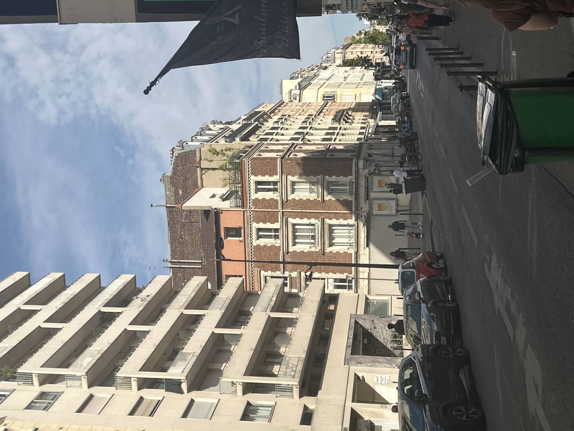 The image depicts a street scene in Paris, France, likely during the daytime given the bright sunlight casting shadows. The perspective is from a slightly elevated viewpoint, looking down onto the street and buildings.
The primary subjects are the various buildings lining the street, characterized by their Parisian architectural style, featuring multiple stories, balconies, and distinct window designs. The buildings exhibit a mix of brick and lighter-colored facades. In the foreground, a modern building with a unique stepped balcony design stands out.
Along the street, several cars are parked, including a small red car and darker colored sedans. There are also people visible walking on the sidewalk and in the street, some appearing to be commuters or shoppers. Bicycles are also present, some parked and others presumably being ridden. A large green trash bin with a black trash bag overflowing is prominently placed on the right side of the frame. A black flag with white text is visible in the upper left corner.
The sky is partly cloudy, indicating fair weather. The scene is busy with the presence of vehicles, pedestrians, and the overall urban environment, suggesting a typical day in a lively Parisian neighborhood. Some of the visible text on the flag appears to be in French, potentially indicating a business or event.