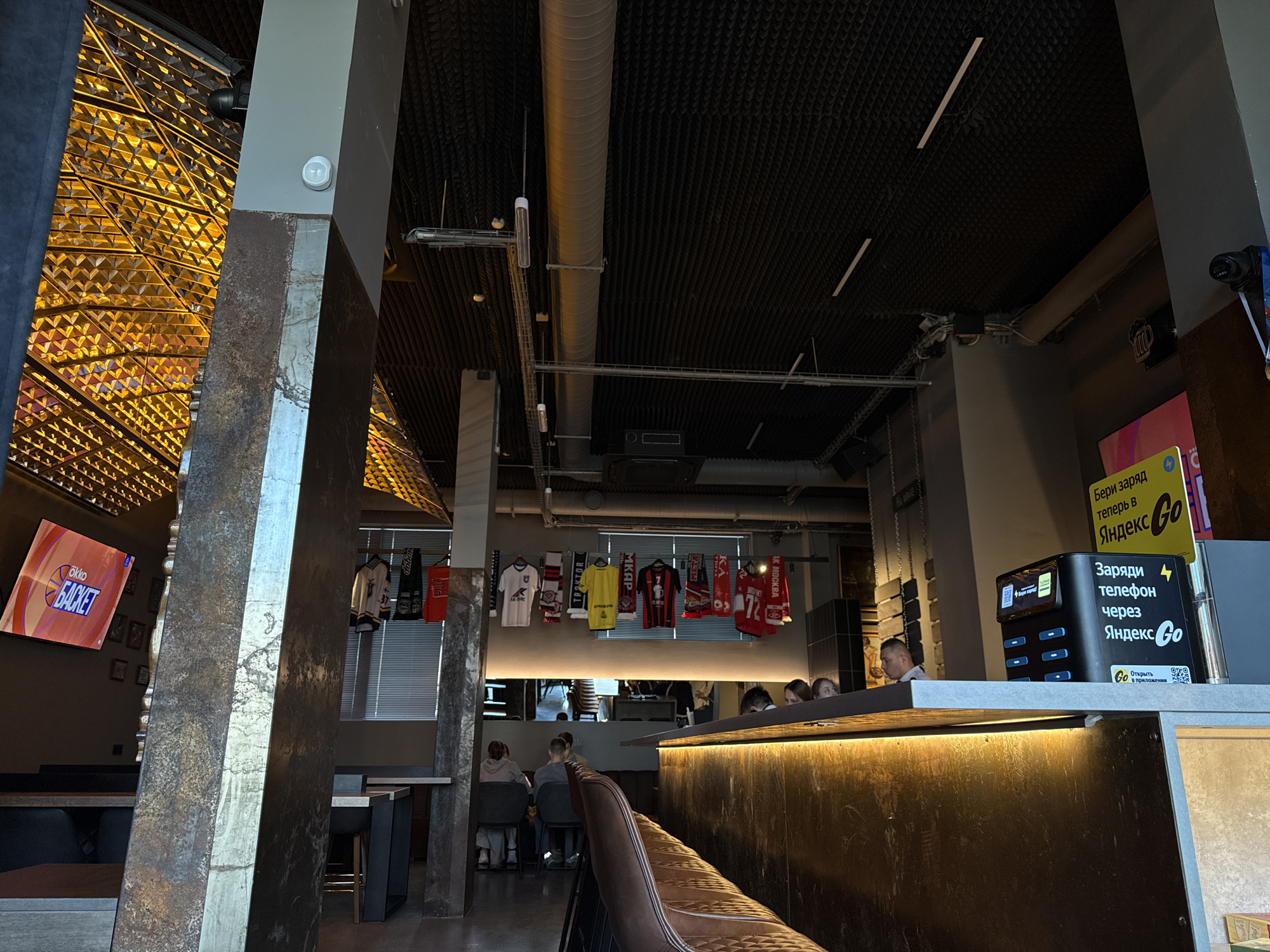 The image shows the interior of a restaurant or bar in Perm, Russia.
Several people are seated at tables and at the bar. Jerseys and scarves are hanging on the wall above the bar. On the bar counter is a mobile phone charging station branded with the "Yandex Go" logo. The TV screen displays a basketball game sponsored by "Okko Basket." The restaurant has a modern, industrial design with exposed ductwork and metal pillars.