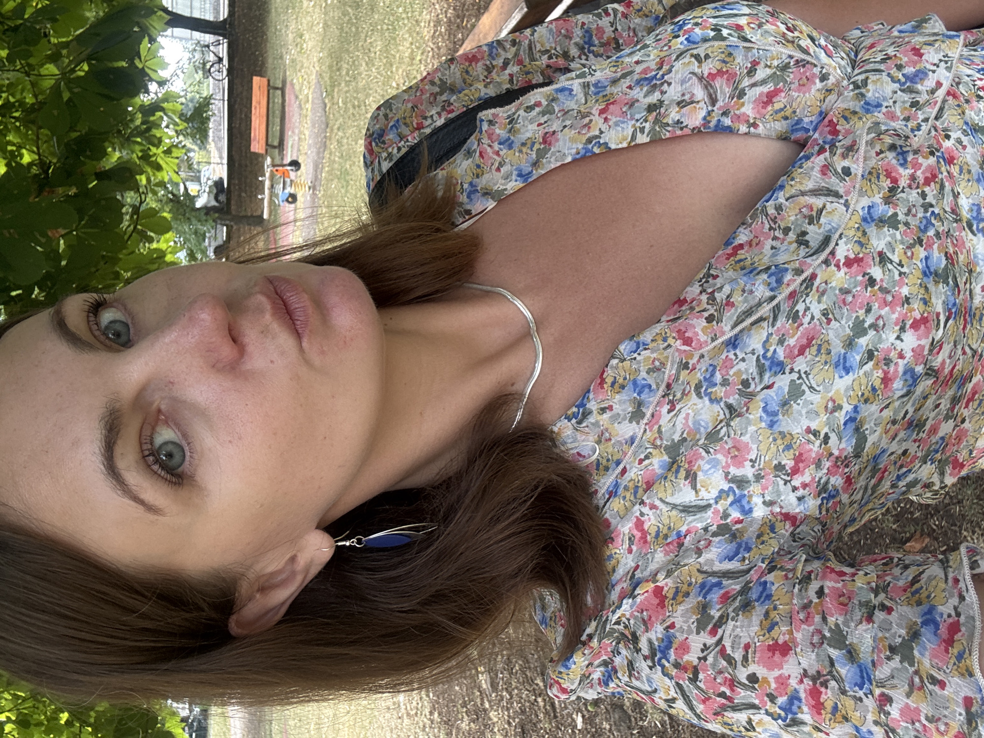 A woman with brown hair and blue-green eyes takes a selfie outdoors in a park in Divonne-les-Bains, France. She is wearing a floral-print dress and a silver necklace, and a blue earring hangs from her left ear. Her mouth is pursed as if she is about to kiss. In the background, trees, a park bench, and a playground are visible.