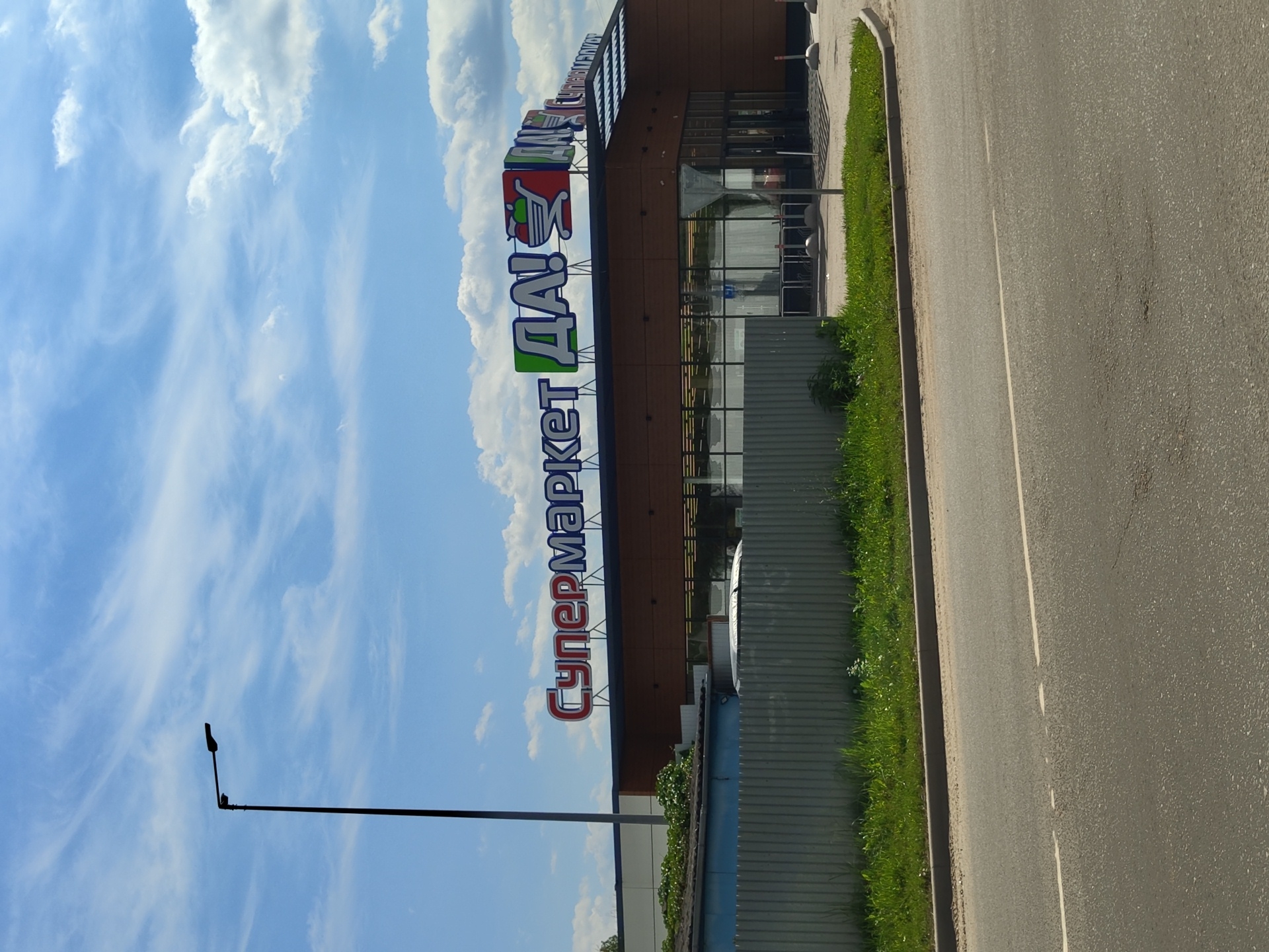 The image shows a street-level view of a supermarket building with a large sign that reads "СУПЕРМАРКЕТ ДА!" in red and green letters. The supermarket has a modern facade with large glass windows and a wooden exterior. In front of the supermarket, there is a metal fence and a strip of green grass. The street in the foreground is paved and has white lane markings. A lamppost is visible to the left of the building, silhouetted against a blue sky with scattered clouds. The scene appears to be in Zhukovsky, Russia, as indicated by the Cyrillic signage. There are no people visible in the image.