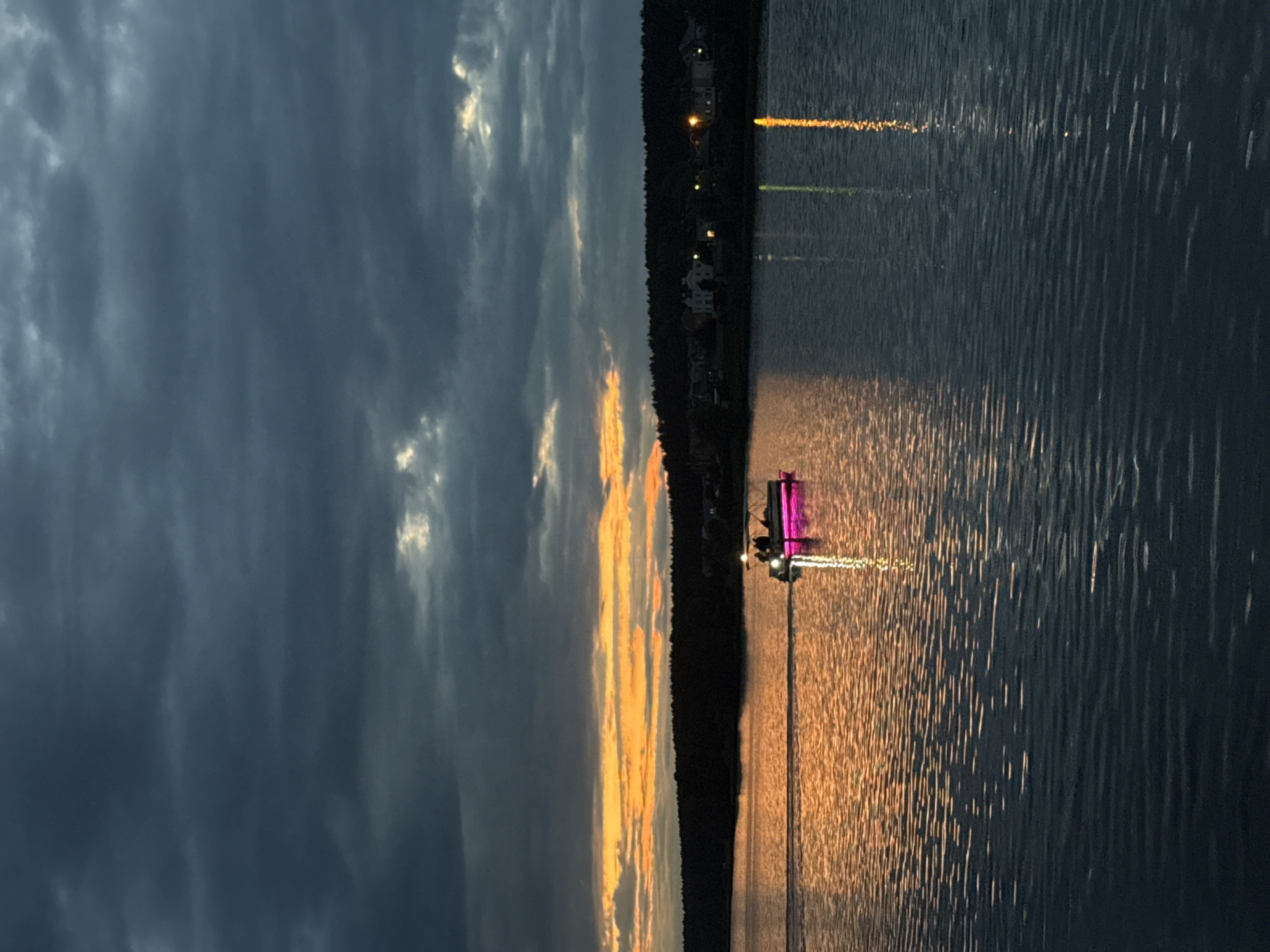 The image shows a pontoon boat with pink LED lighting on the water at dusk. The boat has at least three people on board. The sky is cloudy with a band of orange and yellow sunset visible on the horizon, casting a golden reflection on the water. A dark forest borders the far shore, with lights from houses visible in the distance. The water is rippled, and the reflection of the sunset and the boat's lights are visible. This scene is located on the Votkinsk Reservoir, near Votkinsk, Russia, known for its scenic beauty and recreational activities.