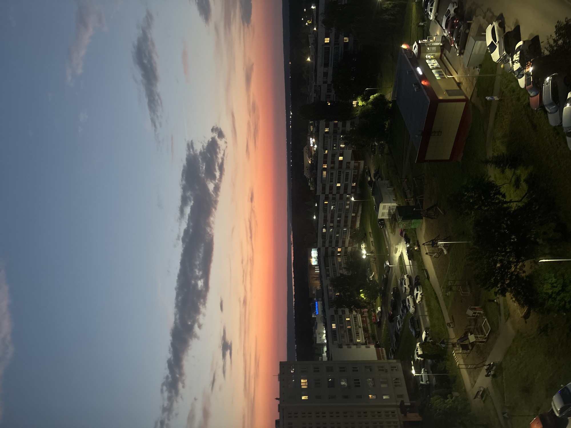 The image depicts an aerial view of a residential area at dusk in Naberezhnye Chelny, Russia. The sky transitions from a light blue at the top to a vibrant orange and pink gradient near the horizon, indicating the setting sun. Clouds are scattered across the sky. In the foreground, tall apartment buildings are visible, some with illuminated windows. A playground with swings and climbing structures is situated between the buildings. Several cars are parked in a lot adjacent to one of the buildings and along a street. A small commercial building with a yellow and red facade, possibly a store or cafe, is also present, with cars parked outside it. Trees and grassy areas are interspersed throughout the scene. The overall impression is of a typical urban environment at the end of the day.