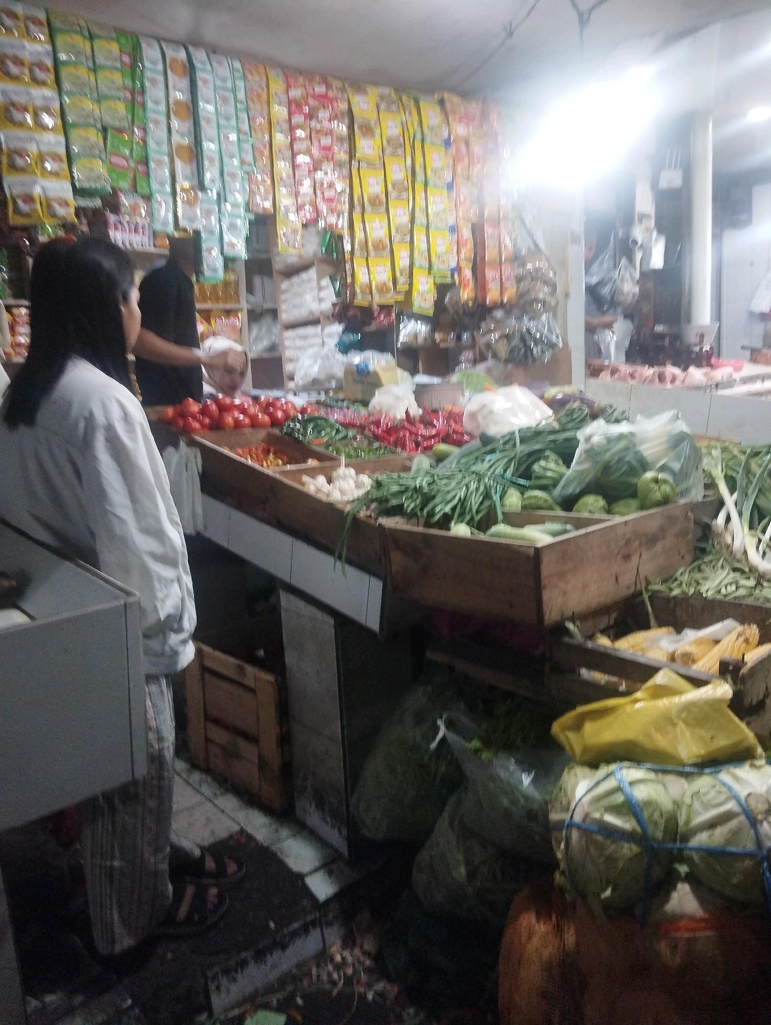 The image depicts a bustling market stall in Malang, Indonesia, likely an outdoor or semi-outdoor setting given the lighting and displays. The stall is laden with a variety of fresh produce, including tomatoes, chilies, leafy greens, cucumbers, and cabbages. Packaged goods, possibly snacks or spices, are neatly hung on the wall above the produce. In the foreground, a person wearing a light-colored shirt and patterned pants stands near a counter. Another individual, partially obscured, appears to be a vendor behind the display of vegetables. The scene is vibrant and suggests a typical Indonesian market environment.