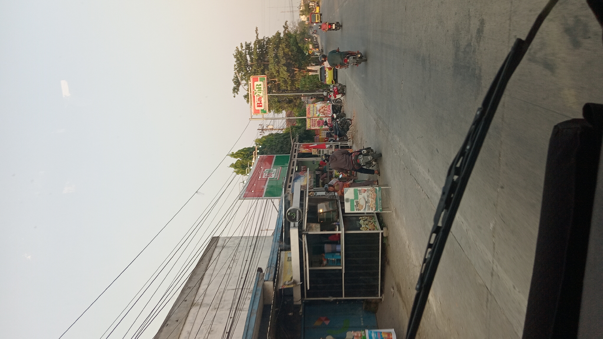 This image depicts a street scene in Kunduran, Indonesia, likely during daylight hours given the bright sky. In the foreground, the windshield wipers and part of a vehicle's dashboard are visible, indicating the photo was taken from inside a car. The road is paved and appears to be a two-lane street with some cracks and markings.
To the left of the road, a building with a light-colored facade and a blue awning stretches along the street. In front of this building, a small food stall is set up with a black counter and various items displayed, including what appear to be drinks and food. Several people are present at or near the food stall; one person is standing at the counter, and another is seated on a motorcycle parked nearby. A sign on an easel is also visible in front of the stall, displaying images of food.
Further down the street, on the right side, a prominent sign with the word "Bazar" in red and white lettering against a green background is mounted on a pole. This suggests the presence of a marketplace or commercial area. Several other signs with promotional text are also visible. Multiple motorcycles are parked along the roadside, and several people are riding motorcycles on the street, moving in the same direction as the implied vehicle taking the photo.
The environment includes some trees behind the "Bazar" sign, contributing to a somewhat urban or suburban feel. Overhead, power lines crisscross the sky, a common feature in many Indonesian cities. The overall atmosphere is one of everyday street life, with commercial activity and transportation visible. The sky is clear and light, suggesting a possibly warm day.