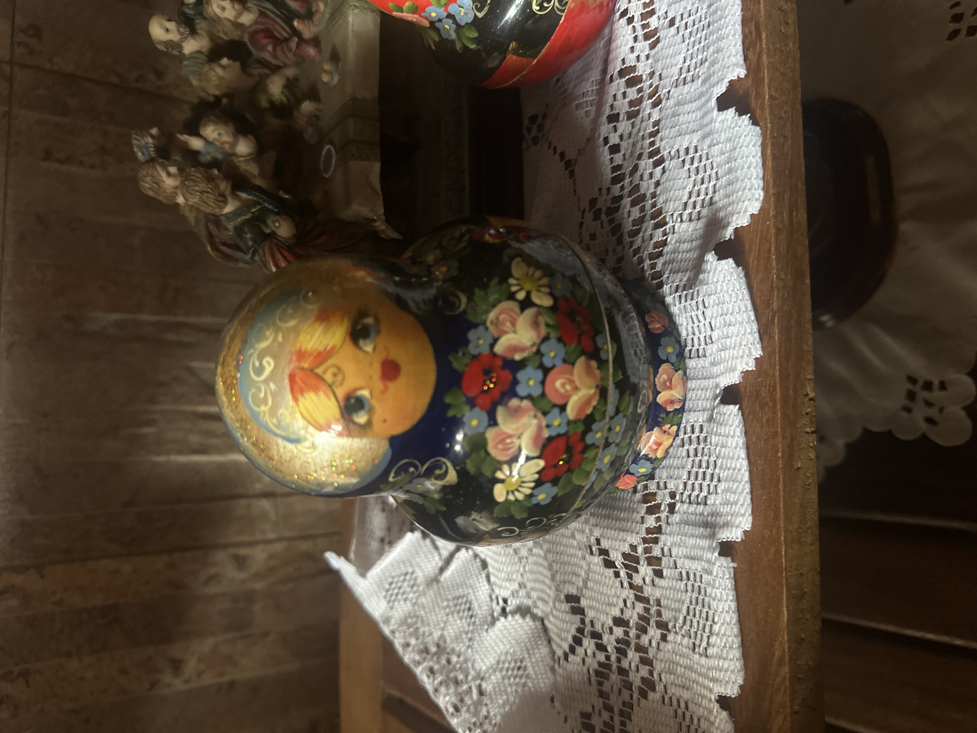 A close-up shot focuses on a decorated Russian nesting doll, known as a Matryoshka doll, positioned on a wooden shelf. The doll is primarily dark blue with a golden headpiece, adorned with a painted face featuring blue eyes, red lips, and rosy cheeks. The body of the doll is intricately decorated with a pattern of colorful flowers, including pink roses, red poppies, and small blue blossoms. White lace doily lies beneath the doll and another Matryoshka doll, partially visible on the right, is red and black. In the background, a collection of small figurines depicting a religious scene, possibly "The Last Supper," is arranged on a table draped with a patterned cloth. The backdrop consists of a textured, vertical wooden paneling. The scene is illuminated by ambient lighting, casting subtle shadows. The precise location in El Maco, Venezuela is not determinable from the provided image.