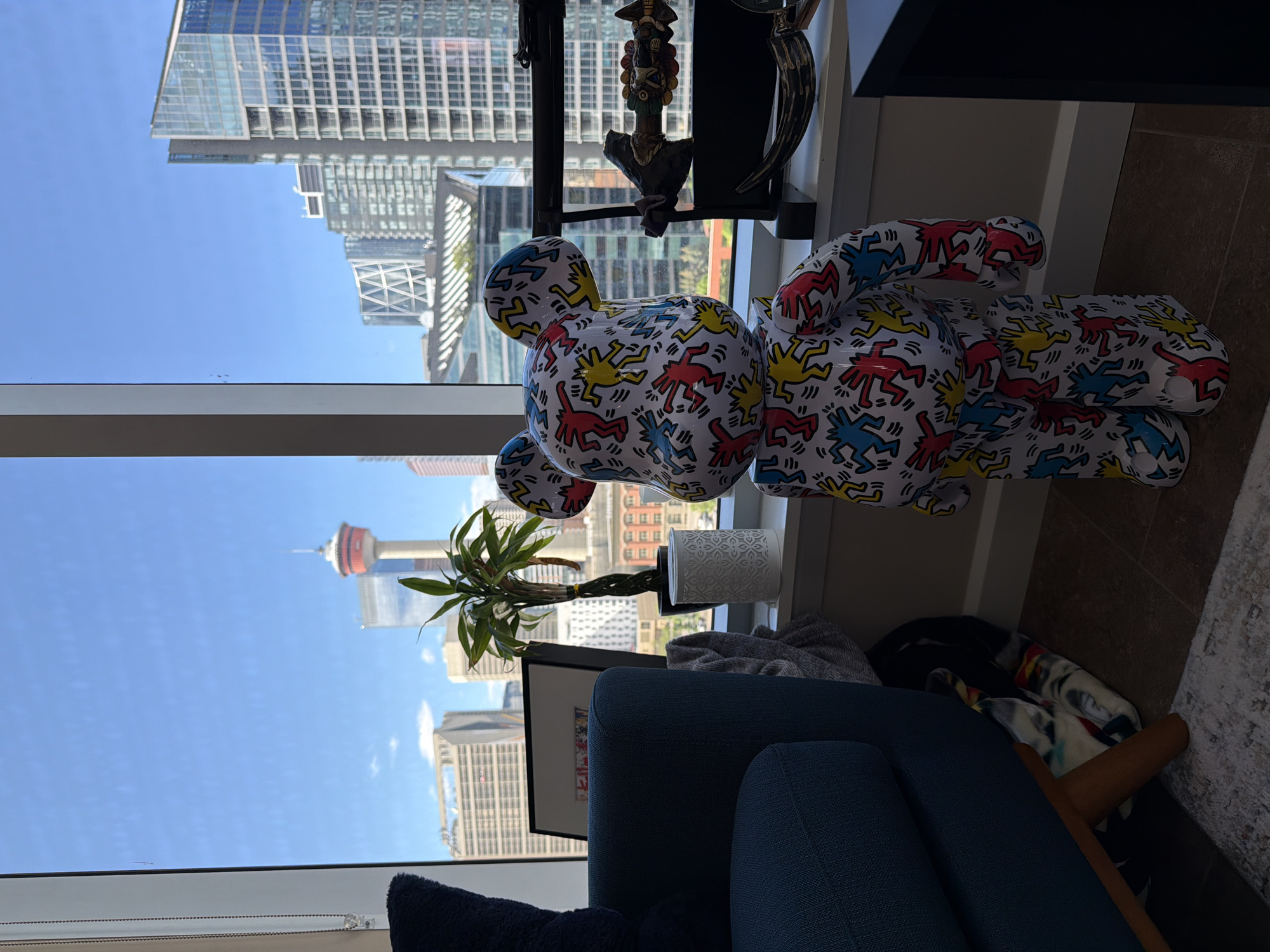 The image depicts an indoor scene with a large window offering a view of the Calgary, Canada skyline. Prominently featured in the foreground is a large Bearbrick toy, decorated with Keith Haring-style figures in red, yellow, and blue. To the left of the Bearbrick, a portion of a blue armchair is visible. On the windowsill behind the Bearbrick, a small potted plant and a white decorative container are placed. The window reveals several city buildings, including the Calgary Tower, and other modern skyscrapers. The overall impression is a well-lit room with a cityscape view.