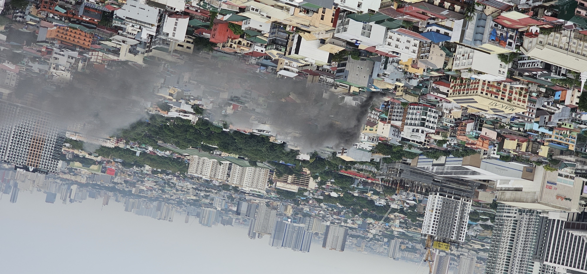 This is an aerial photograph of a dense urban area in Makati, Philippines, characterized by a mix of high-rise buildings and lower-rise residential and commercial structures. A plume of dark smoke is rising from a cluster of buildings in the middle ground, indicating a fire. The smoke is thick and appears to be spreading, obscuring some of the surrounding structures. The sky is overcast, contributing to a hazy atmosphere that diminishes visibility of distant buildings. In the foreground and mid-ground, a variety of buildings with different colors and architectural styles are visible, showcasing the dense urban fabric of the city. Some buildings are under construction, indicated by cranes. The overall scene depicts an active urban environment with an ongoing fire incident.