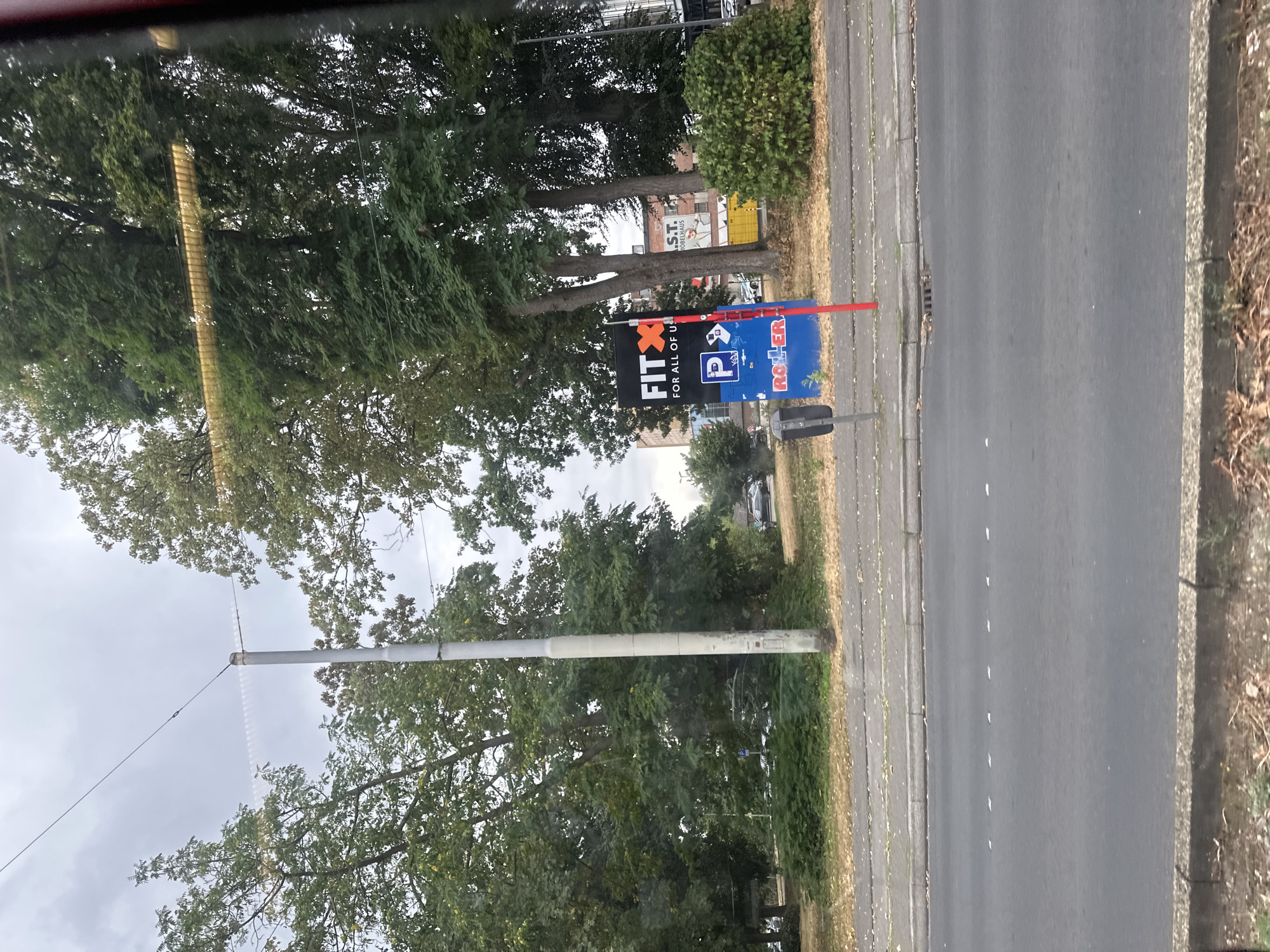 The image shows a street scene in Krefeld, Germany, on a cloudy day. In the foreground, a street is visible with road markings and a curb. Behind the street, there is a sidewalk and a grassy area with a pole and a sign. The sign is black with "FITX" in large white letters and "FOR ALL OF US" below it. There is also a blue parking sign with a white "P" and a red sign below it with "ROLLER" written in white letters. Trees line the background, and buildings are visible between them. Some cars are parked in the distance. The scene appears to be a commercial area.