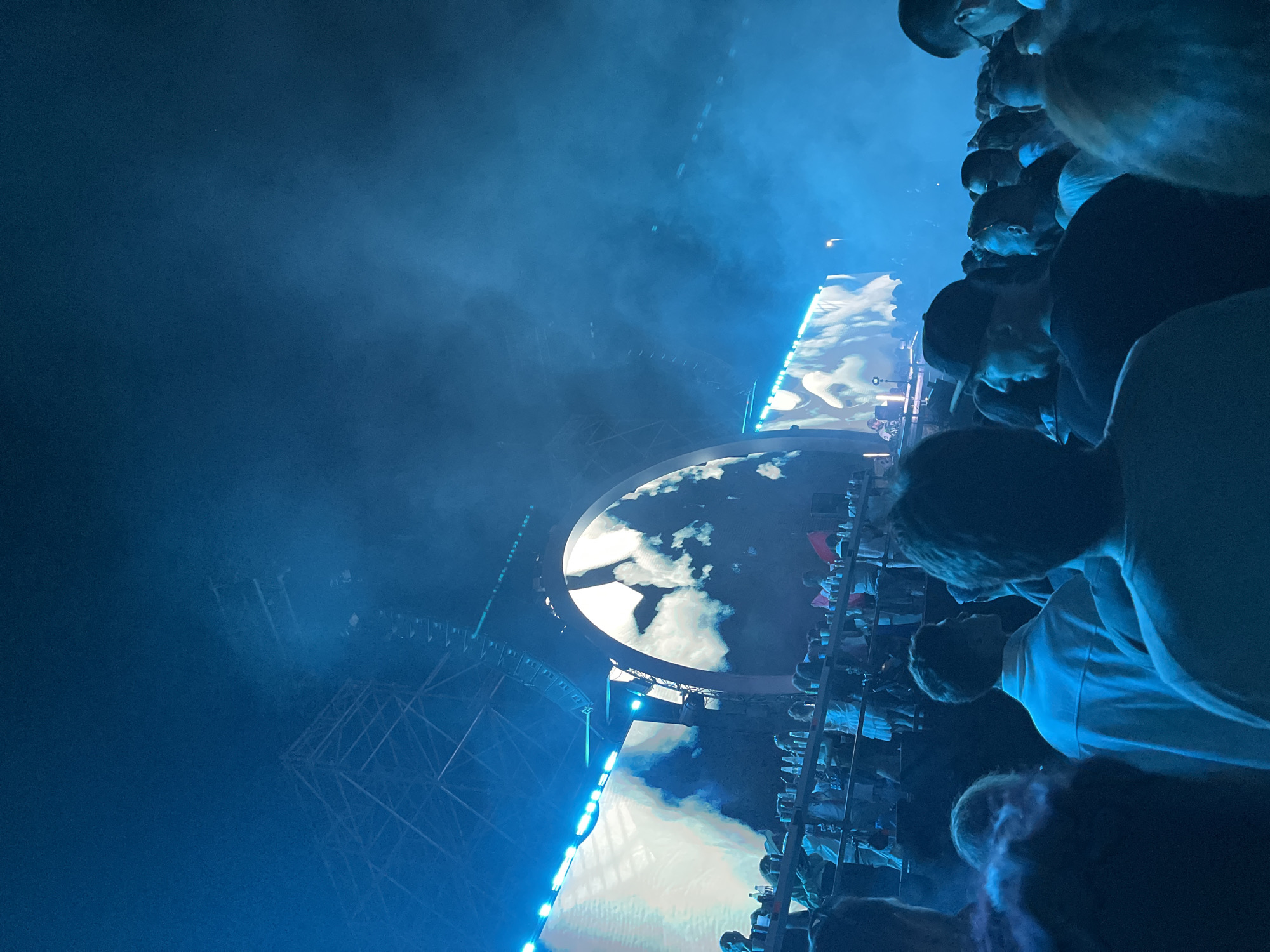 The image depicts a nighttime outdoor music concert. A large crowd of people is gathered, facing a stage area. Dominating the background is a massive, arched LED screen displaying a swirling, cloud-like visual. To the left of the arched screen, another large LED screen shows a lighter, more abstract image. Above the screens, intricate scaffolding and lighting rigs are visible against the dark sky, partially obscured by haze or smoke. The crowd is densely packed, with individuals silhouetted by the stage lighting. Many people are wearing casual clothing. The atmosphere is illuminated by vibrant blue stage lights, creating a moody and energetic ambiance. The location is identified as Sofia, Bulgaria.