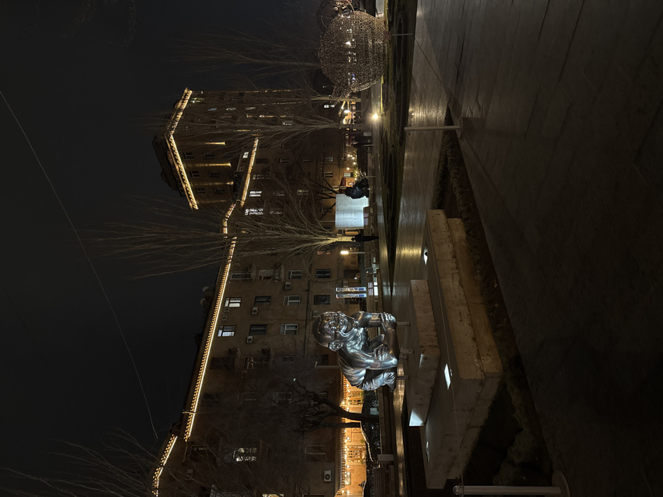 The image captures Charles Aznavour Square in Yerevan, Armenia, at night. In the foreground, a prominent, polished metal sculpture of a crawling baby, identified as "My and Our Baby," is situated on a multi-tiered concrete base illuminated by embedded ground lights. To the mid-ground right, a large, glowing, wire-frame sculpture resembling a teapot is visible. The background consists of multi-story buildings, their rooflines and some windows illuminated by decorative string lights and internal lighting. One building exhibits a taller section with vertical accent lighting. Bare deciduous trees are scattered throughout the scene. Paved walkways and landscaped areas are illuminated by numerous ground-level lights. Several indistinct human figures are present in the mid-ground, primarily near the buildings and other sculptures. The sky is dark and clear.