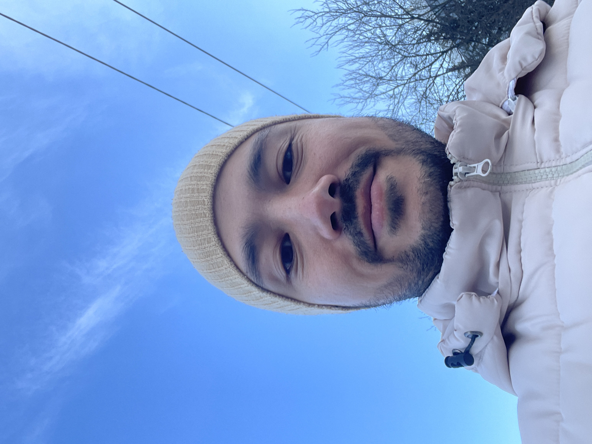 The scene is an outdoor, low-angle selfie captured in Izhevsk, Russia. An adult male subject, appearing to be in his 20s to 40s with dark eyes and a short beard and mustache, occupies the majority of the frame, looking directly into the camera with a neutral to slightly smiling expression. He is wearing a beige knitted beanie and a light beige or pale pink padded winter jacket with a visible zipper. The background features a bright blue sky with sparse white cirrus clouds. Two parallel overhead power lines are visible in the upper right quadrant. Bare deciduous tree branches are present in the lower right quadrant, suggesting a winter or early spring environment.