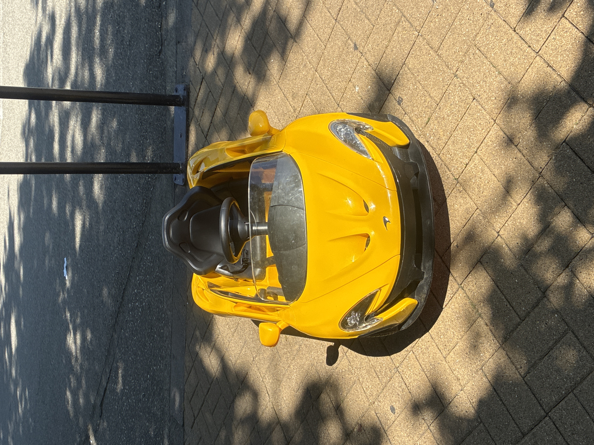 A bright yellow toy McLaren is parked on a cobblestone sidewalk. The toy car has a black plastic seat and steering wheel. The car is facing forward and is angled slightly to the left. Shadows from trees are cast across the car and the sidewalk. In the background, two black poles are visible, and beyond them is a gray asphalt surface. There are no people or animals in the image. The scene appears to be outdoors on a sunny day. This scene is not specifically located in the city of Sochi, Russia; it is a generic outdoor scene.