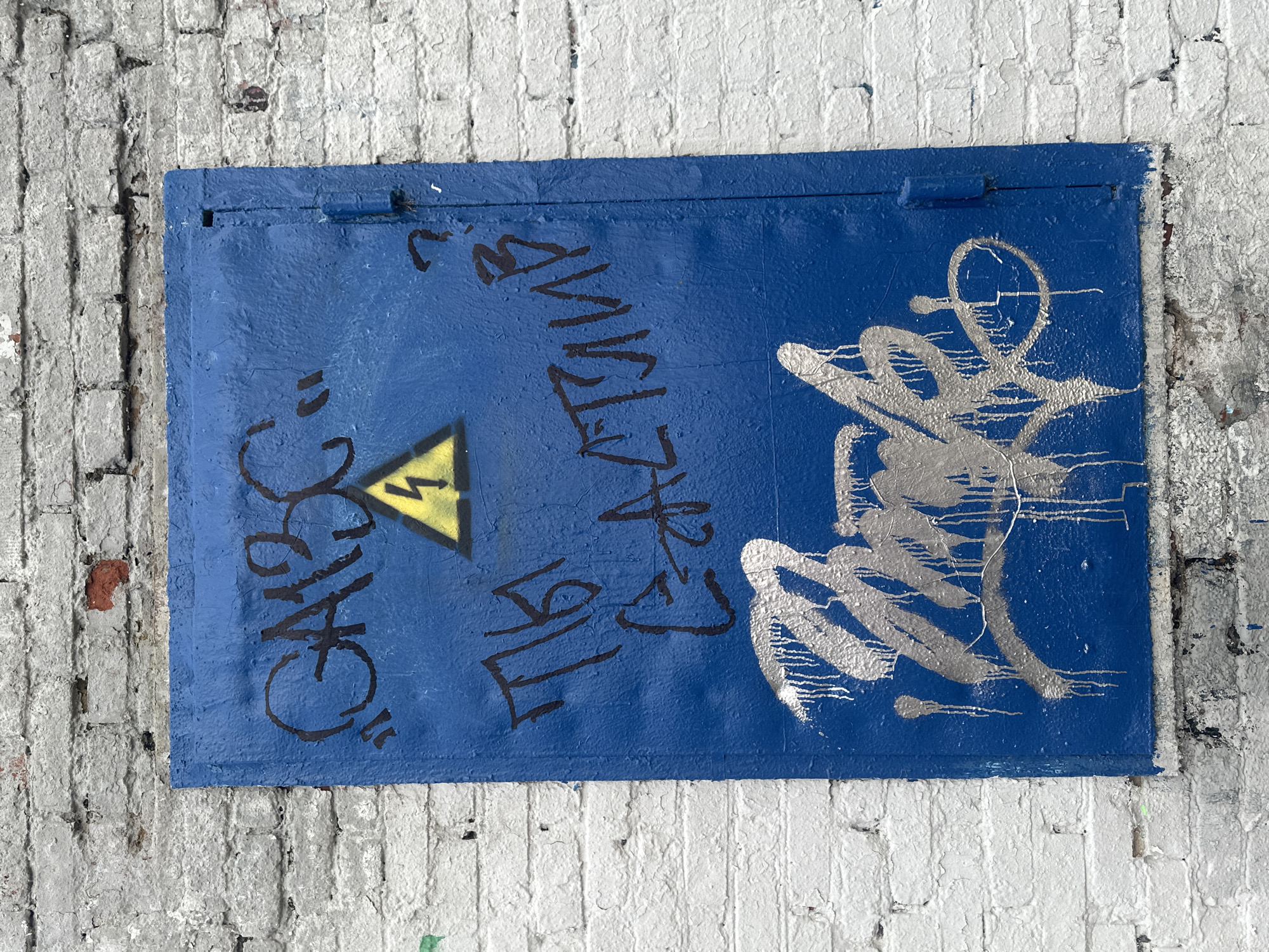 A blue metal door, likely a utility access panel, is mounted on a white-painted brick wall. The door has graffiti, including the word "CHAS" in black, a yellow electrical hazard triangle, and illegible silver spray paint. The wall shows signs of age and weathering. The location is Moscow, Russia.