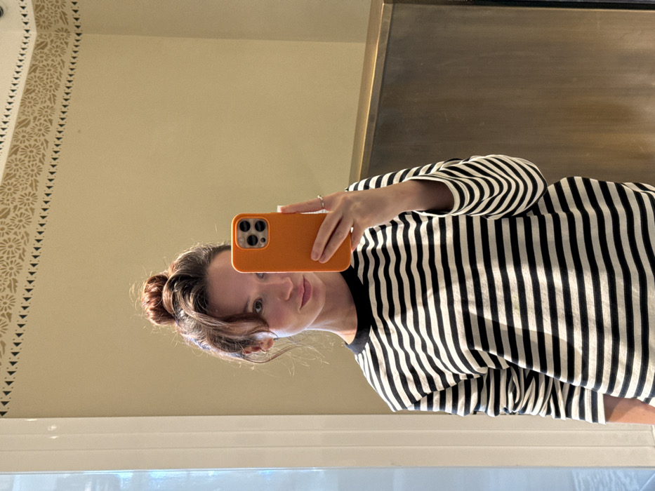 A single female individual is depicted taking a mirror selfie indoors. She has reddish-brown hair styled in a high bun with bangs and is wearing a black and white horizontal striped long-sleeved shirt with a black crew neck. Her right hand holds an orange-cased smartphone, partially obscuring the right side of her face, while a silver-colored ring is visible on her right ring finger. The background consists of a light beige wall featuring a decorative frieze near the ceiling, which includes a band of small black triangles above a band of golden-beige stylized floral motifs. A vertical white architectural trim is present on the far left, and a horizontal panel with a bronze or brass metallic finish is visible on the right. The scene is consistent with an interior room, such as a bathroom or dressing area, located in Tel-Aviv, Israel.