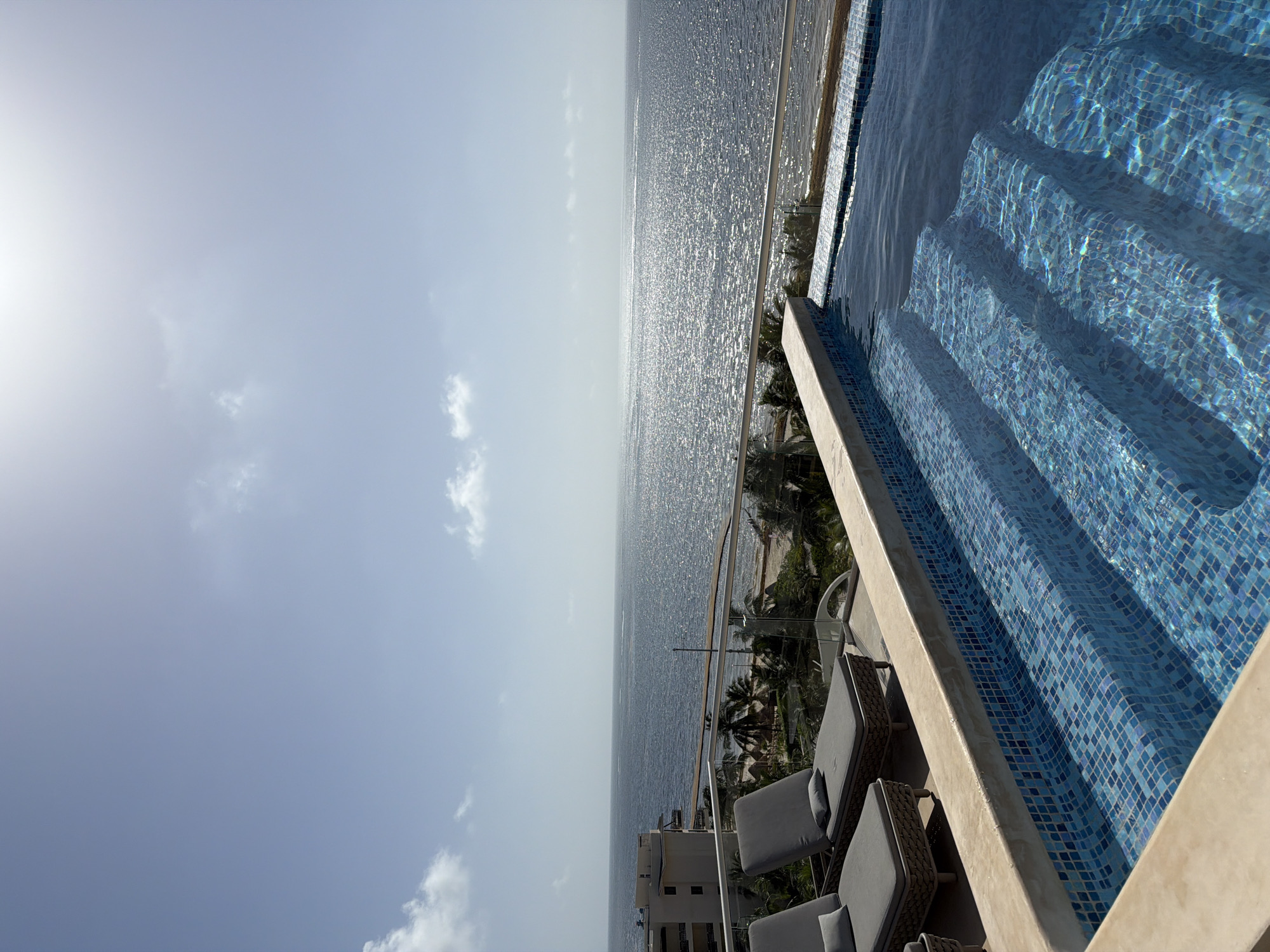 The image shows a rooftop infinity pool overlooking the Caribbean Sea in Puerto Morelos, Mexico. The pool has tiled steps leading down into the clear blue water. Two lounge chairs are visible on the deck next to the pool. In the background, palm trees and a building can be seen along the coastline. The sun is shining brightly, reflecting off the water.