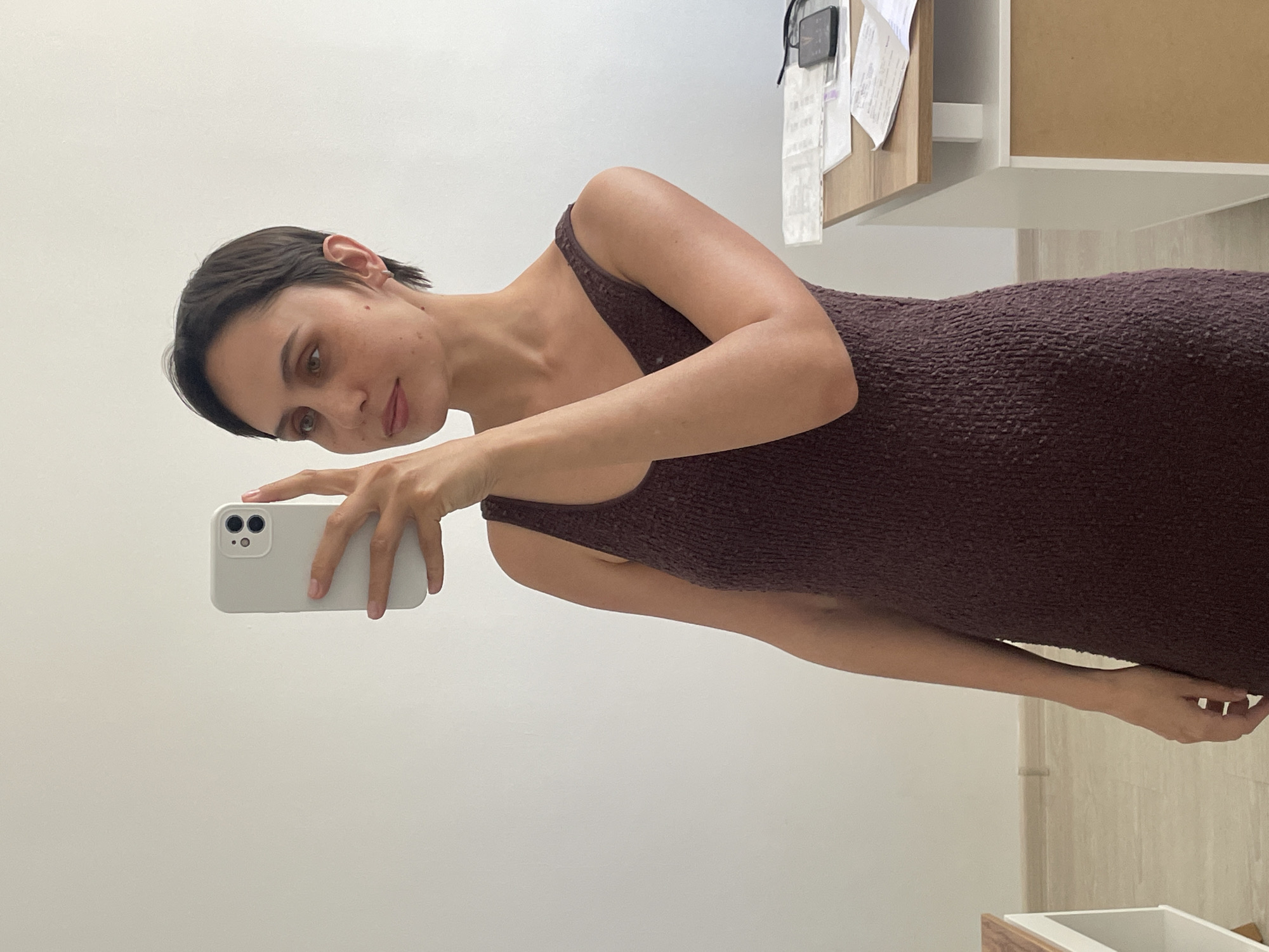 A woman is taking a selfie. She is wearing a brown dress and holding a white smartphone in her left hand. The background is a white wall and part of a wooden cabinet. There is no evidence in the image to suggest a location in Izhevsk, Russia.